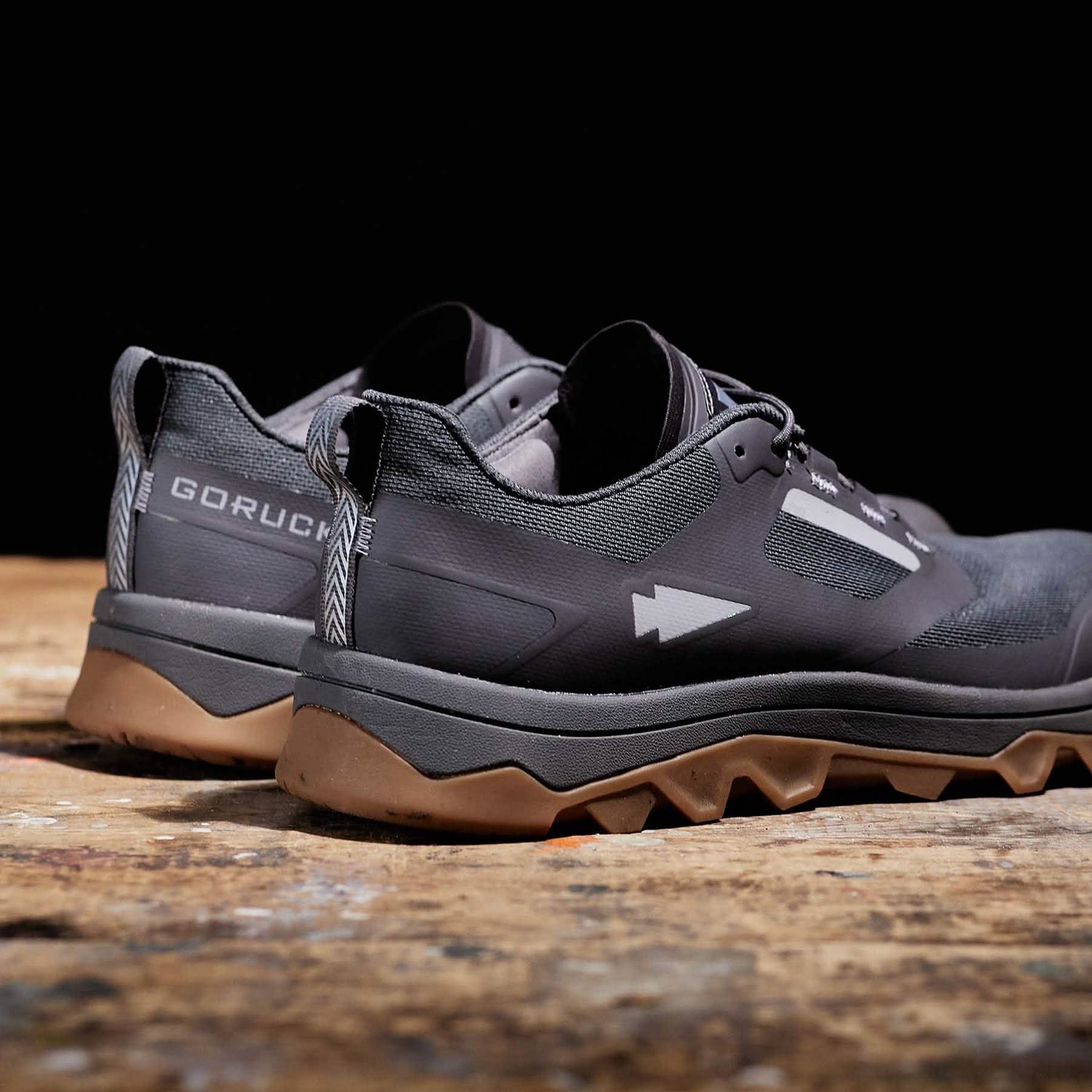 A pair of Mackall trail shoes by GORUCK, in a striking forged iron and chiseled stone color with gum soles, are showcased on a wooden surface. These all-terrain athletic shoes highlight a modern design with branding on the heel and arrow-like graphics along the side, making them ideal for stylishly navigating diverse landscapes.