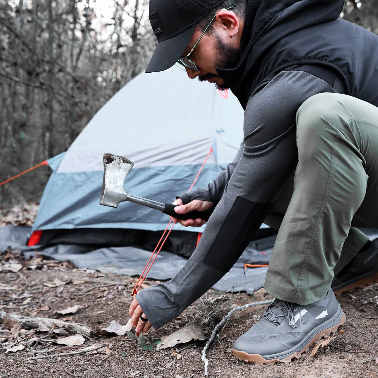 Wearing GORUCK's all-terrain Mackall shoes, a person uses a forged iron axe with a chiseled stone edge to secure a tent stake in the forest. The trail shoes maintain grip on the leaf-covered ground, with their pitched tent visible among towering trees in the background.