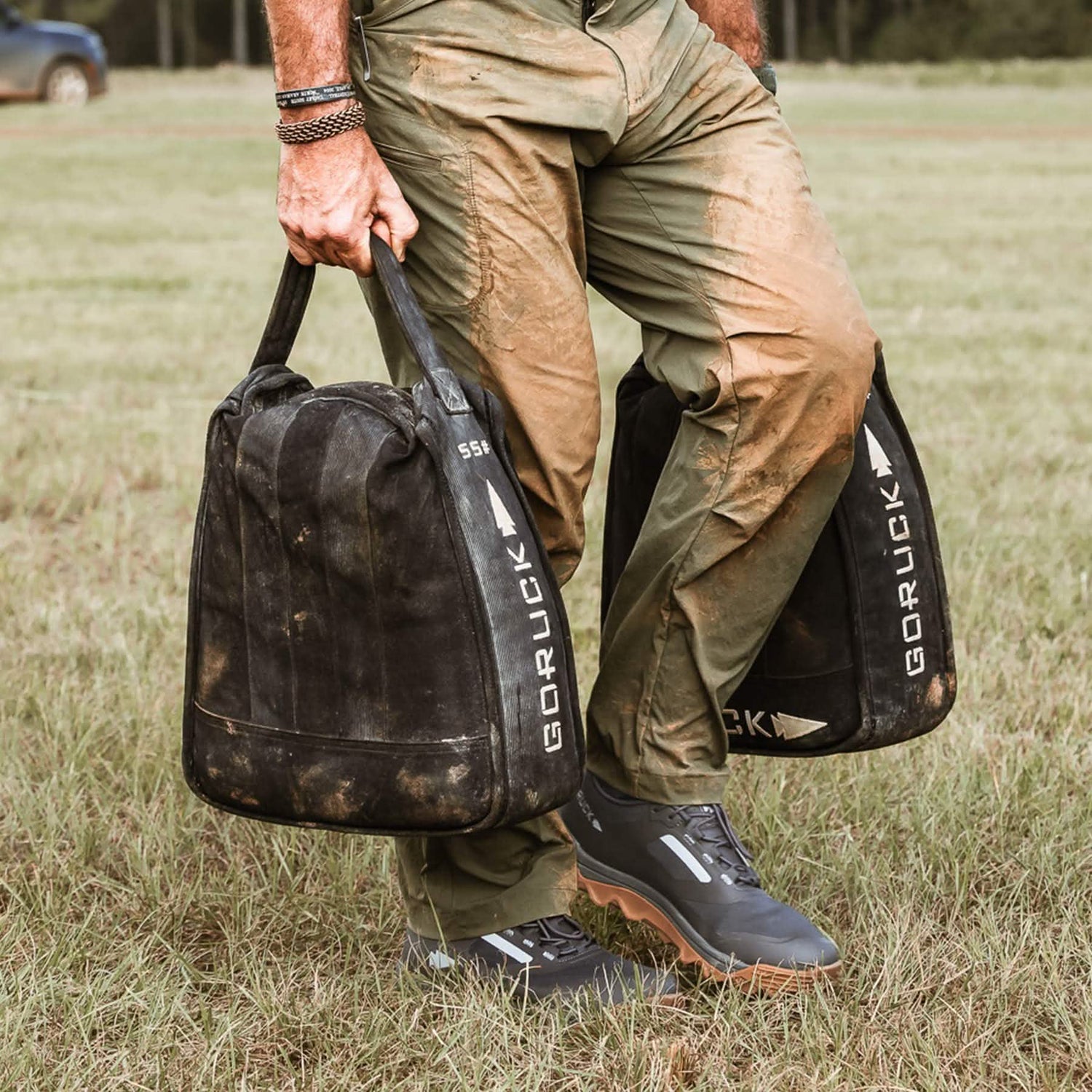 A person carrying two black GORUCK Mackall sandbags over grass, dressed in green pants stained with mud and dark trail shoes. A vehicle and trees can be seen in the background, showcasing their all-terrain adventure.