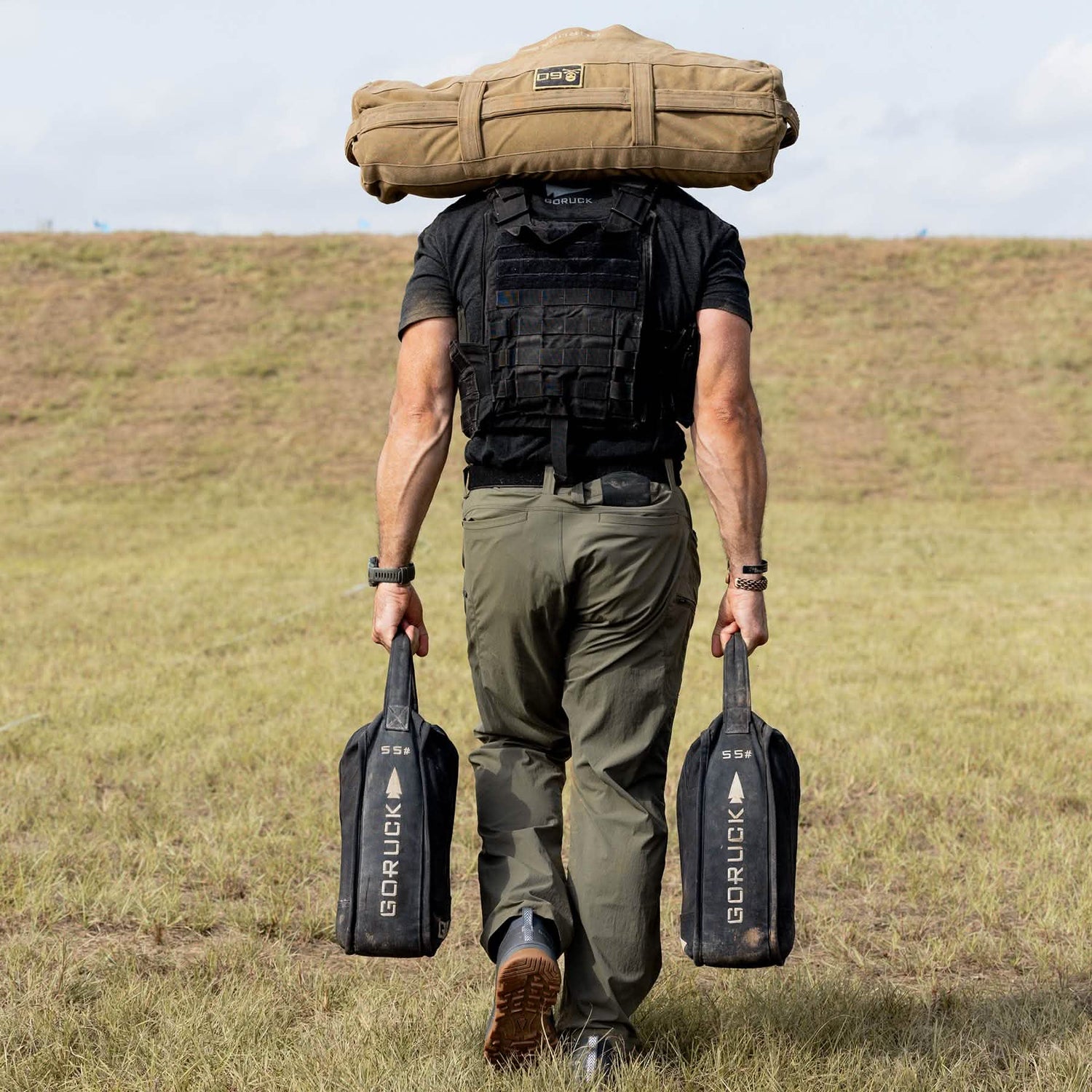 A person strides confidently through the grass, wearing a black tactical vest and Mackall - Forged Iron + Chiseled Stone + Gum rucking shoes by GORUCK. They carry two black sandbags labeled GORUCK and have a large tan backpack slung over their shoulders, as they move under a clear sky dotted with a few clouds.