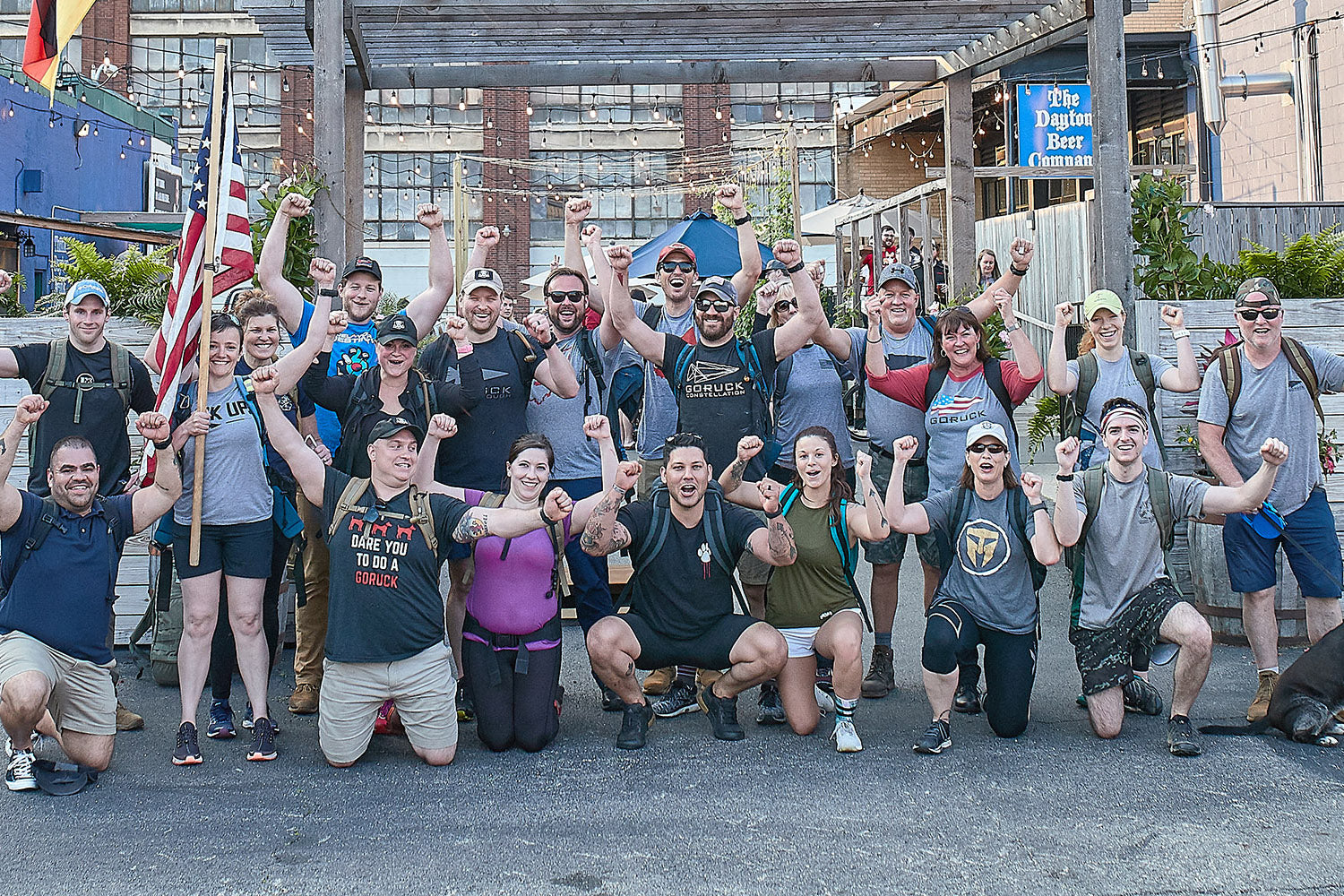 A group of people pose outdoors with raised fists, smiling, some wearing athletic gear and holding an American flag.