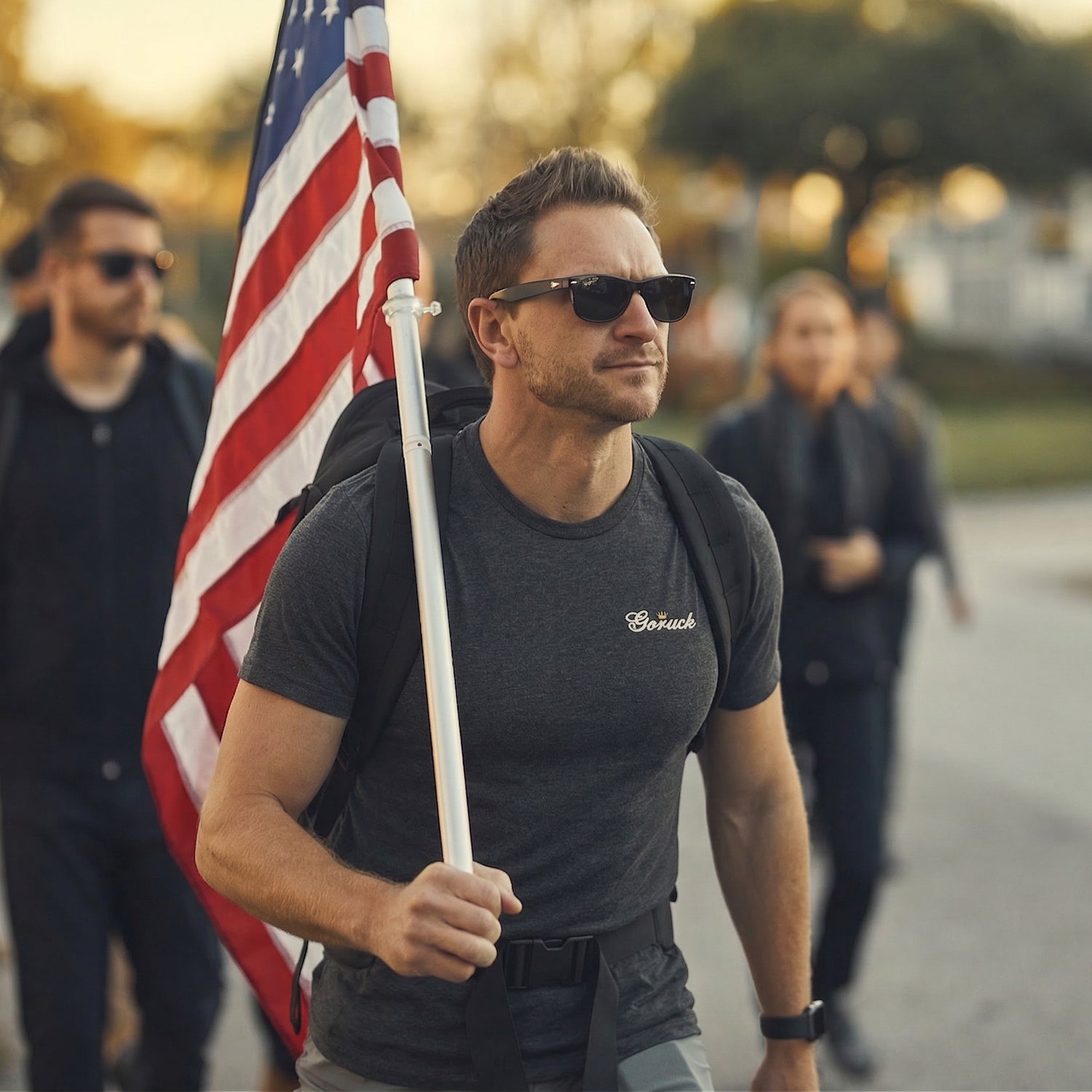 Wearing the GORUCK Bud Tee - Tri-BlendX, a man in sunglasses carries an American flag while walking outdoors with a casually dressed group, combining everyday style and patriotic flair.