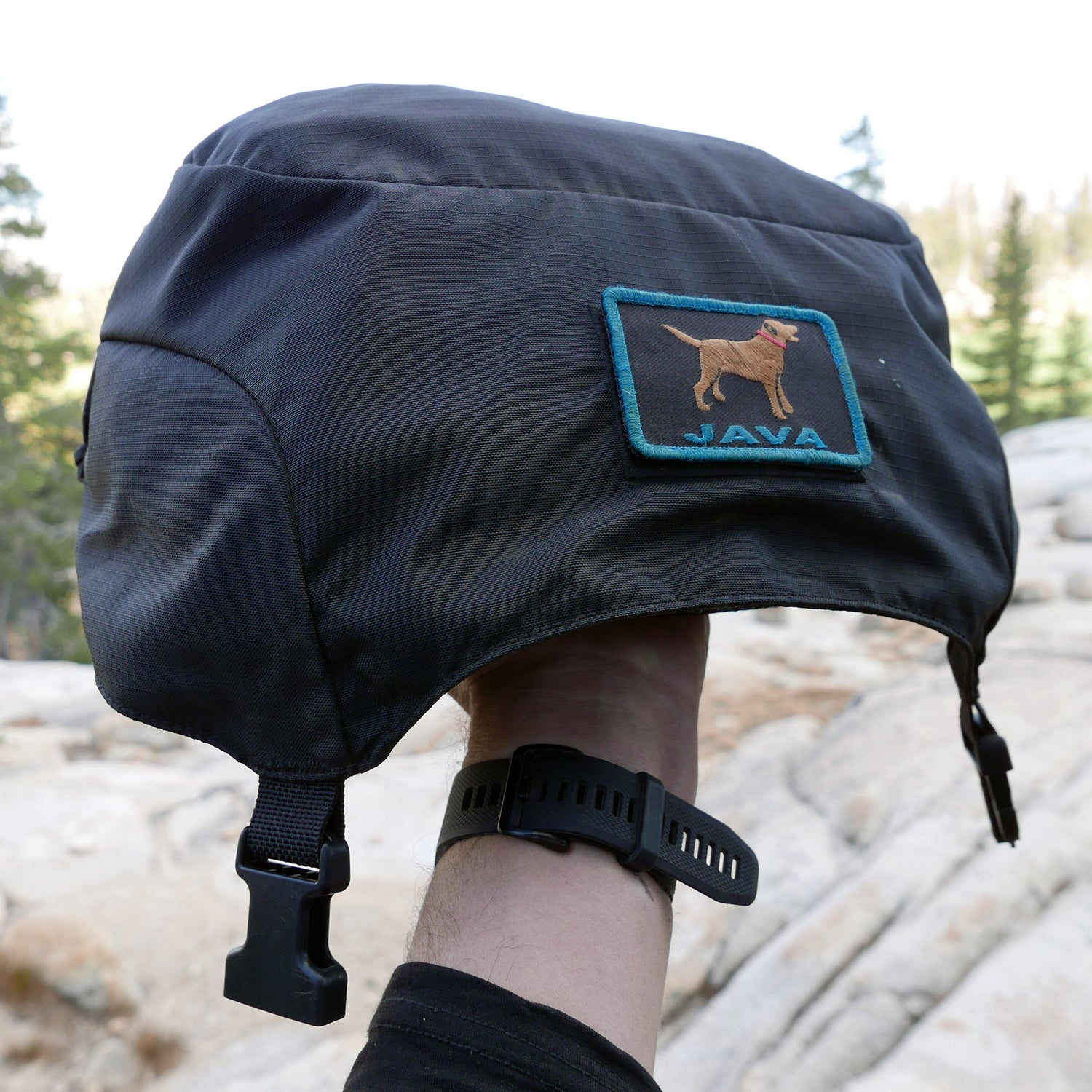 A hand holds a black helmet cover with a patch showing a dog and the word “JAVA” outdoors, next to the rugged GR MONSTER Bundle - PREORDER rucksack from GORUCK.