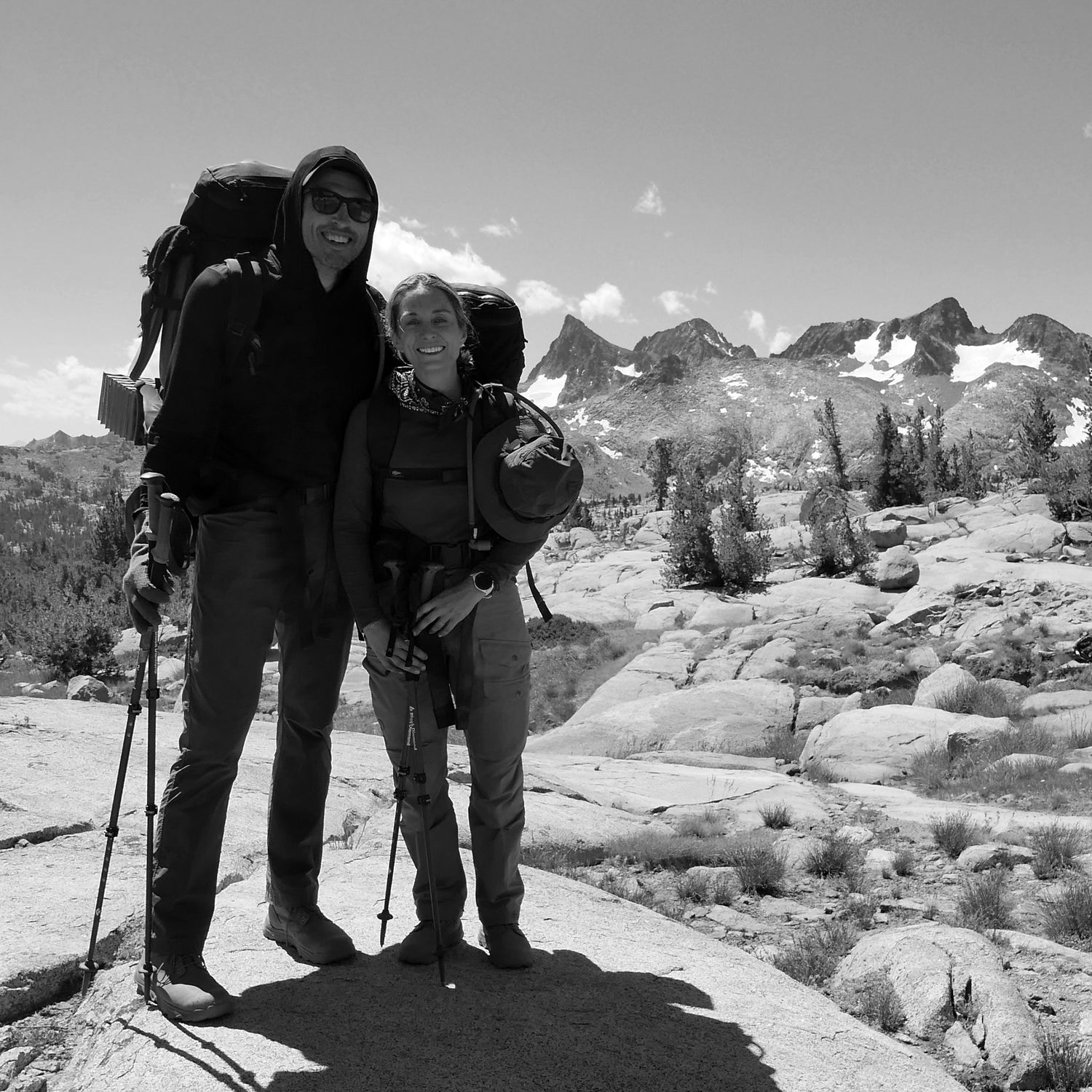Two hikers with rucksacks stand on rocky terrain with snowy mountains in the background, smiling at the camera while showcasing the GR MONSTER Bundle - PREORDER.