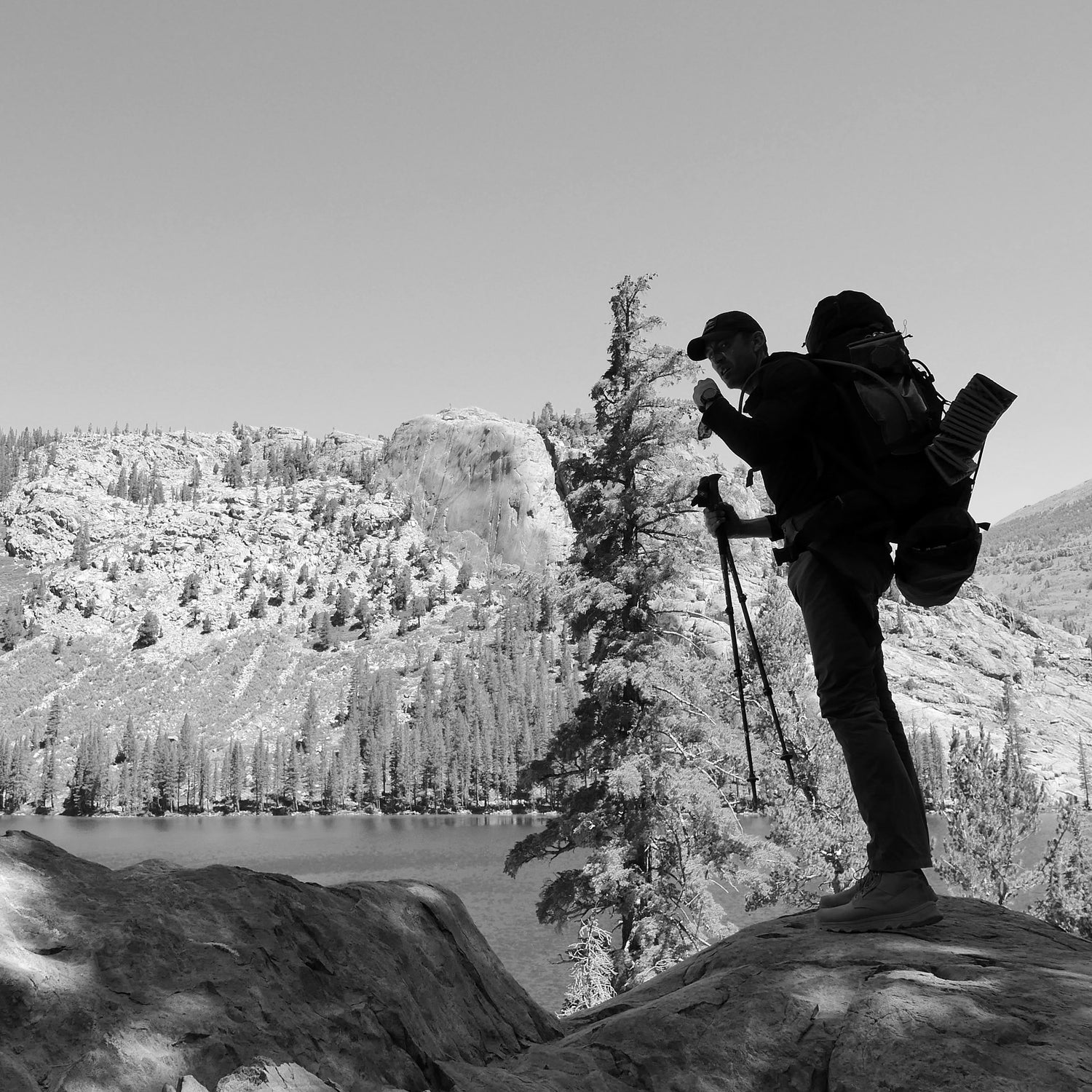 A hiker carrying the GR MONSTER Bundle - PREORDER and a camera tripod stands on a rock overlooking a lake and forested mountains.