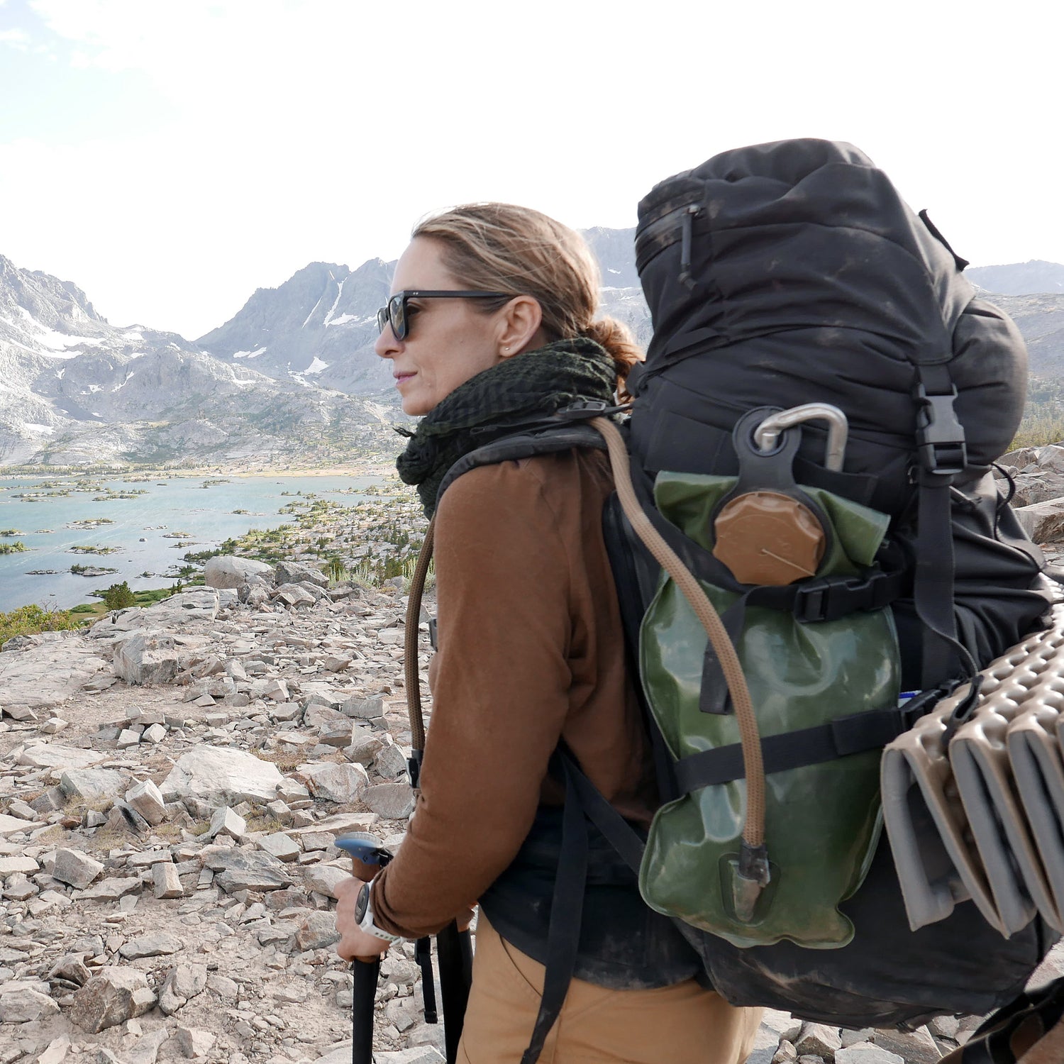 A woman hikes on rocky terrain with the GR MONSTER Bundle - PREORDER rucksack, mountains and a lake in the background.