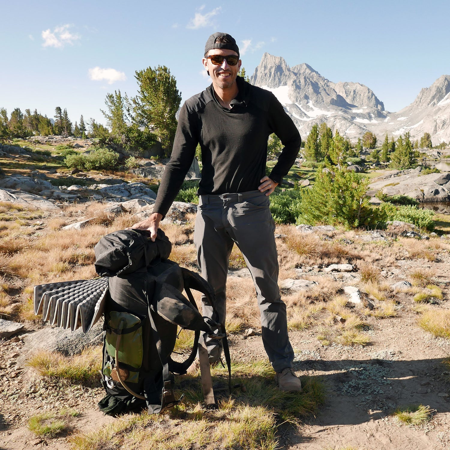 A man in hiking gear stands beside the GR MONSTER Bundle - PREORDER rucksack in a mountain landscape with trees and rocky peaks.