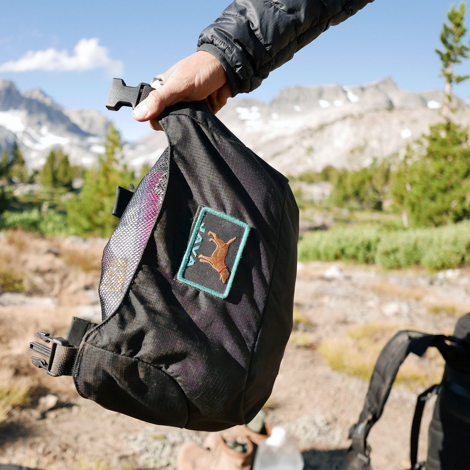 A hand holds a black GR MONSTER Bundle - PREORDER rucksack with a deer patch outdoors, with mountains and trees in the background.
