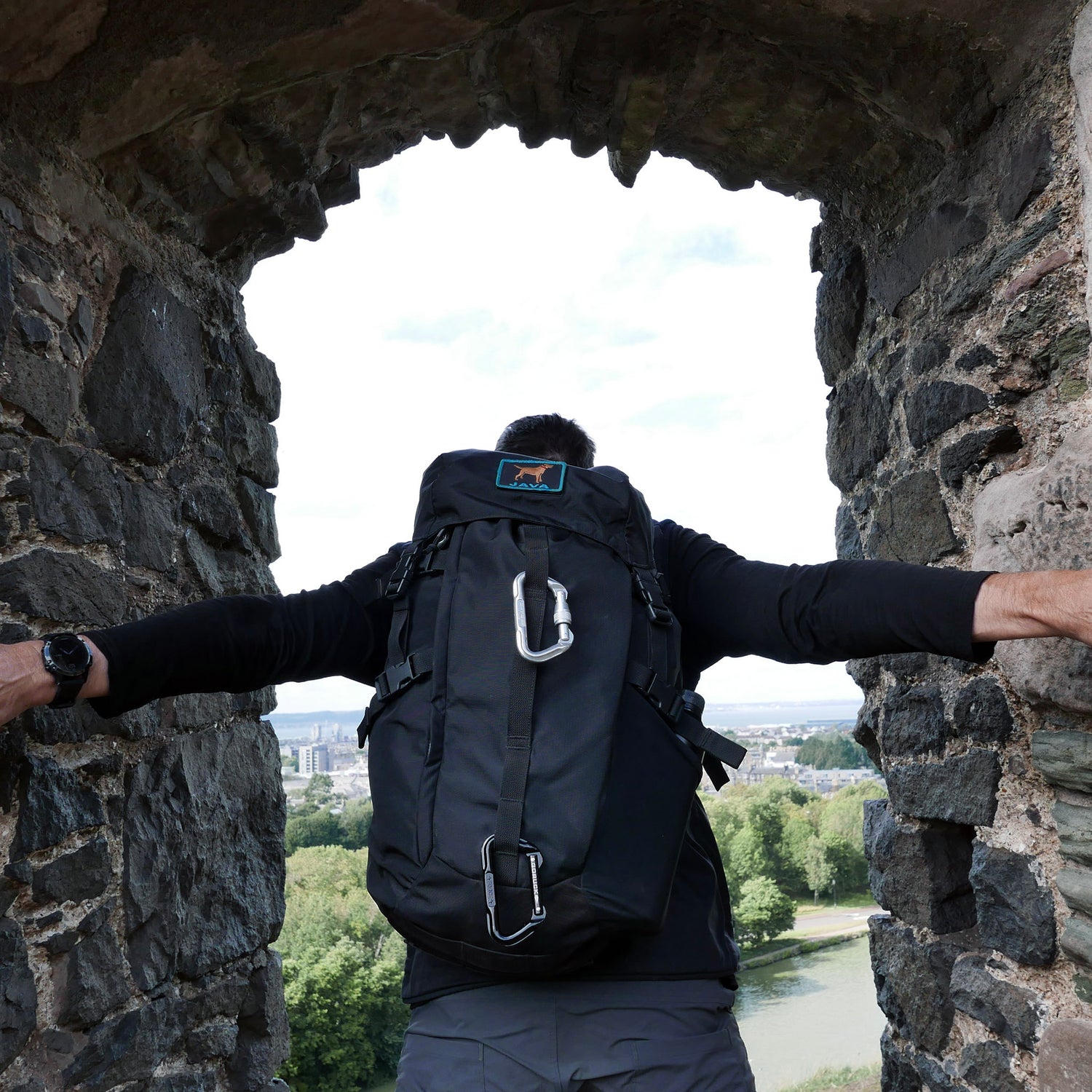 A person with a large black GR MONSTER Rucksack - PREORDER stands in a stone arch, overlooking trees and a distant city.