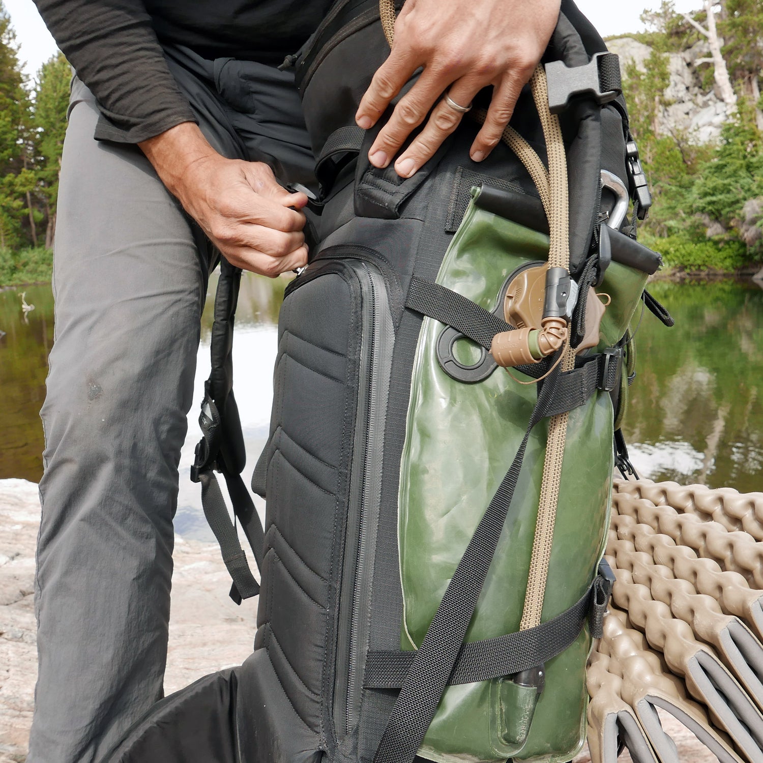 A person securing a GR MONSTER Rucksack - PREORDER in green and black near a body of water in an outdoor setting.
