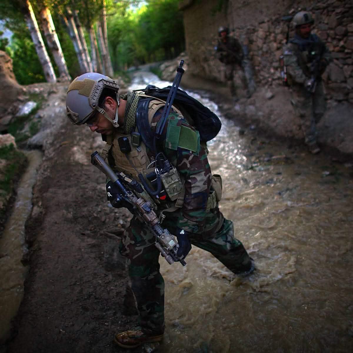 A Special Forces soldier in full gear wades through a shallow stream in a forested area, his durable rucksack keeping essentials dry. In the background, two other soldiers carry their GORUCK GR1 USA - Cordura packs, advancing carefully through the terrain.