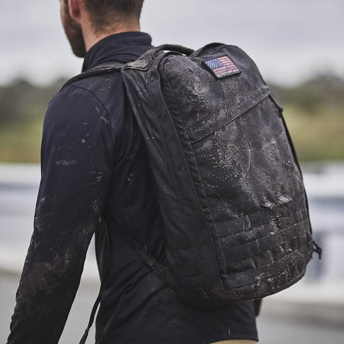 A person carrying a GR1 USA - Cordura backpack by GORUCK, complete with a US flag patch, strides through the outdoors, looking ready for a GORUCK Challenge.