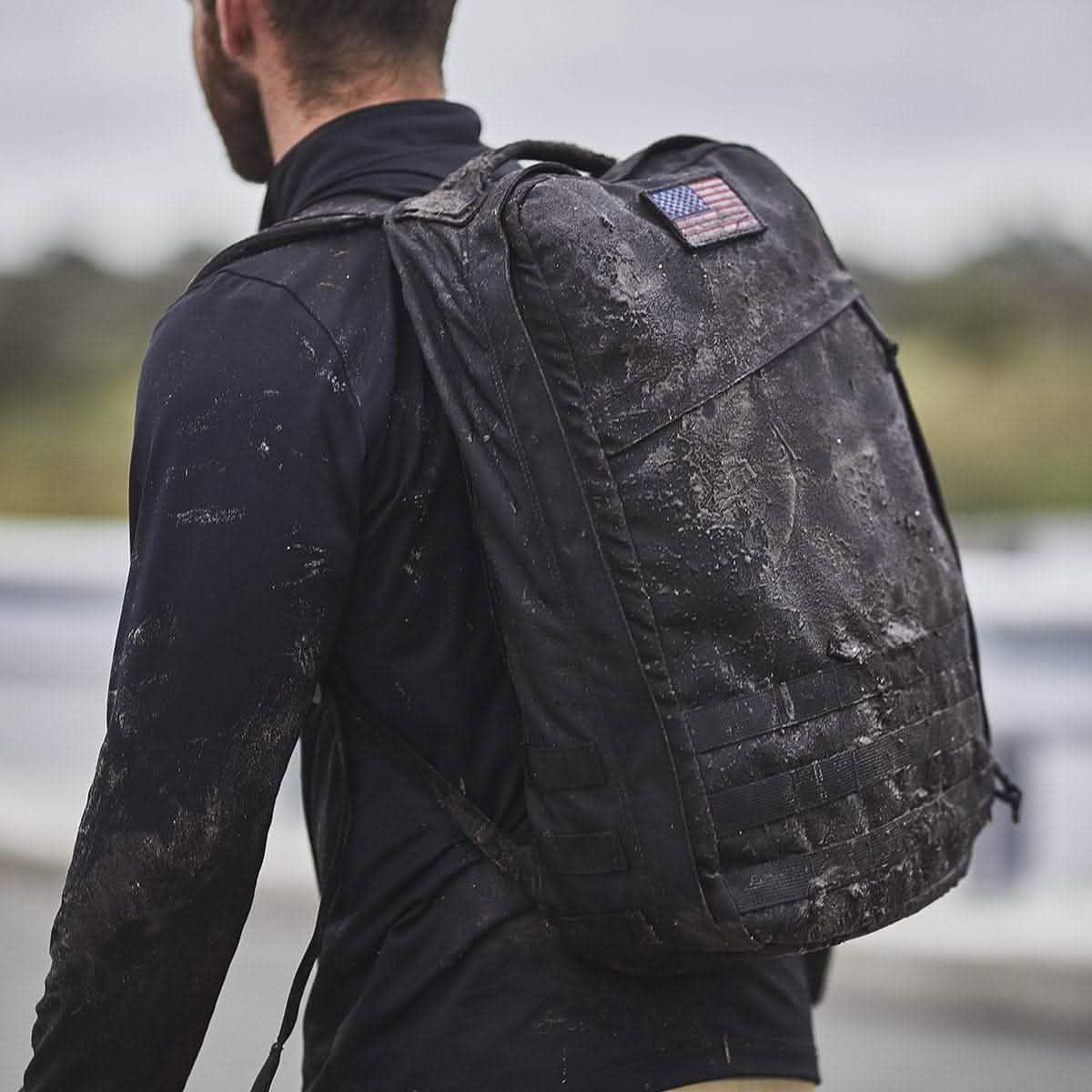 Man wearing muddy black tactical rucksack with American flag patch outdoors
