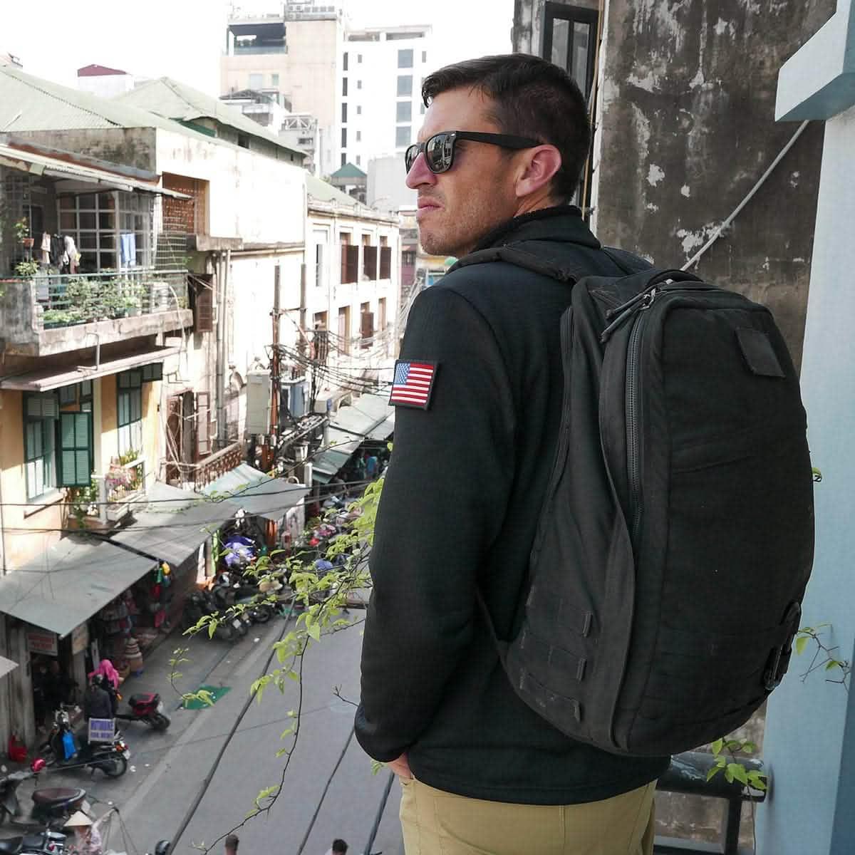Man wearing sunglasses and black tactical backpack overlooking a busy urban street from a balcony