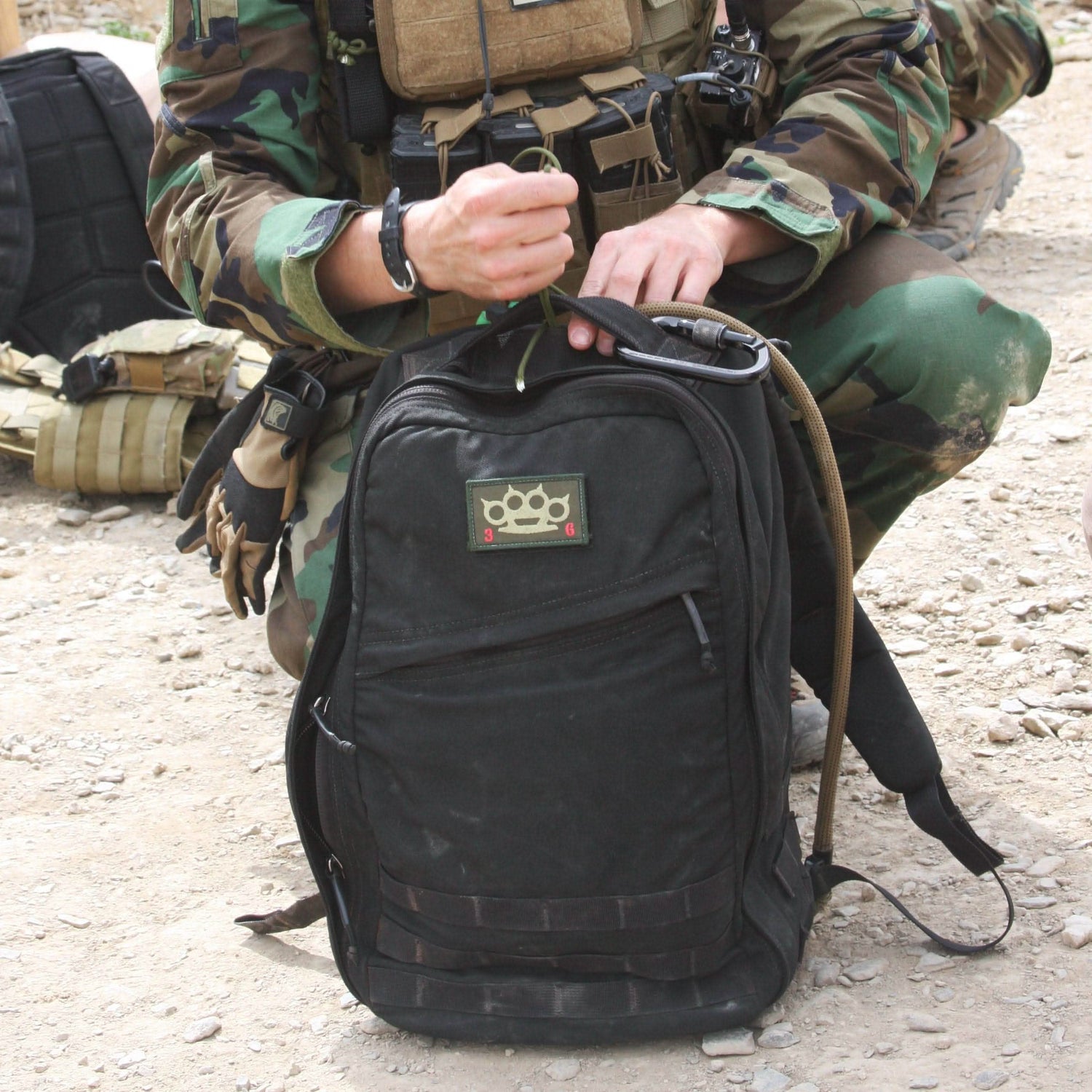 A person in camouflage, embodying the precision of Special Forces, stands outdoors on a dirt surface with a GORUCK GR1 USA - Cordura backpack, proudly displaying an American Manufacturing patch.