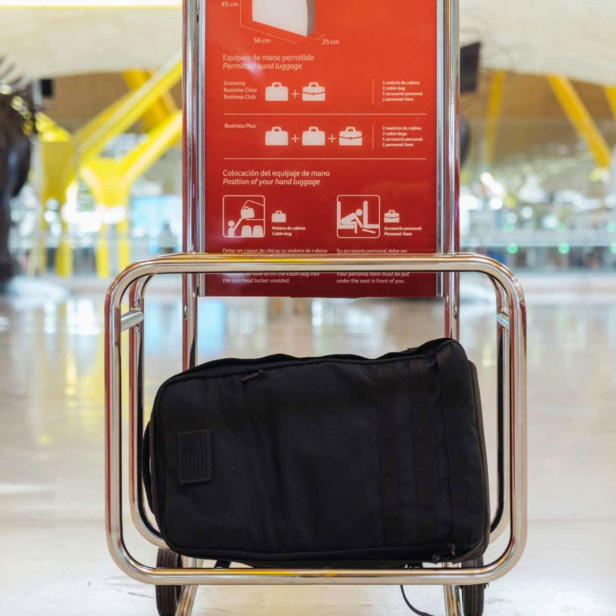 Black GORUCK carry-on bag on airport luggage cart under red hand luggage info sign