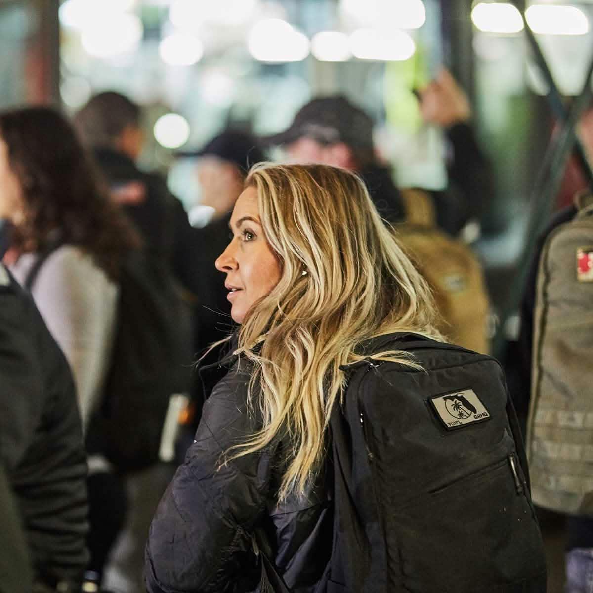 Blonde woman wearing a black jacket and GORUCK backpack in a crowded urban night setting