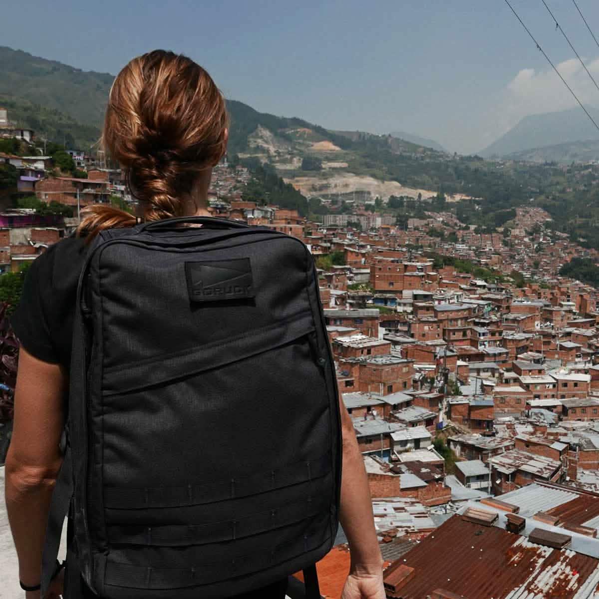 Woman with brown hair in braid wearing black GORUCK backpack overlooking densely packed hillside neighborhood