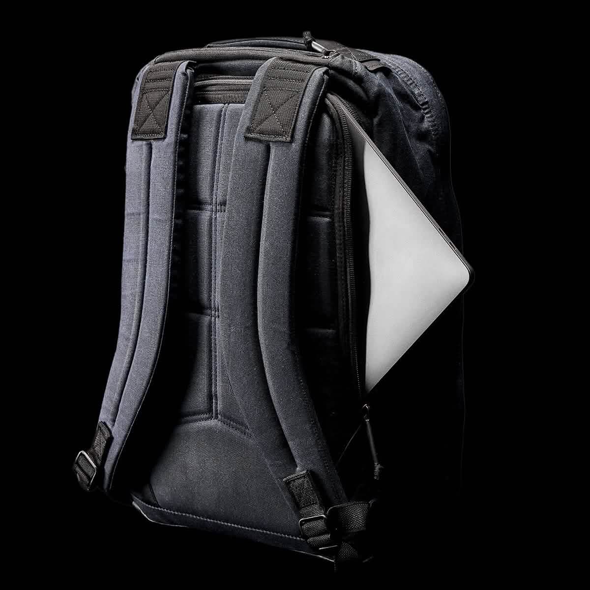 A black GORUCK GR1 USA - Heritage rucksack with padded straps is displayed against a black background. It has a side zipper pocket partially open, revealing the edge of a laptop.