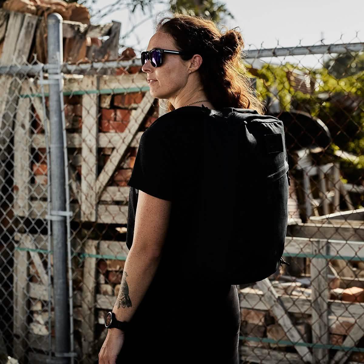 Woman wearing sunglasses and black GORUCK heritage backpack standing near chain-link fence outdoors