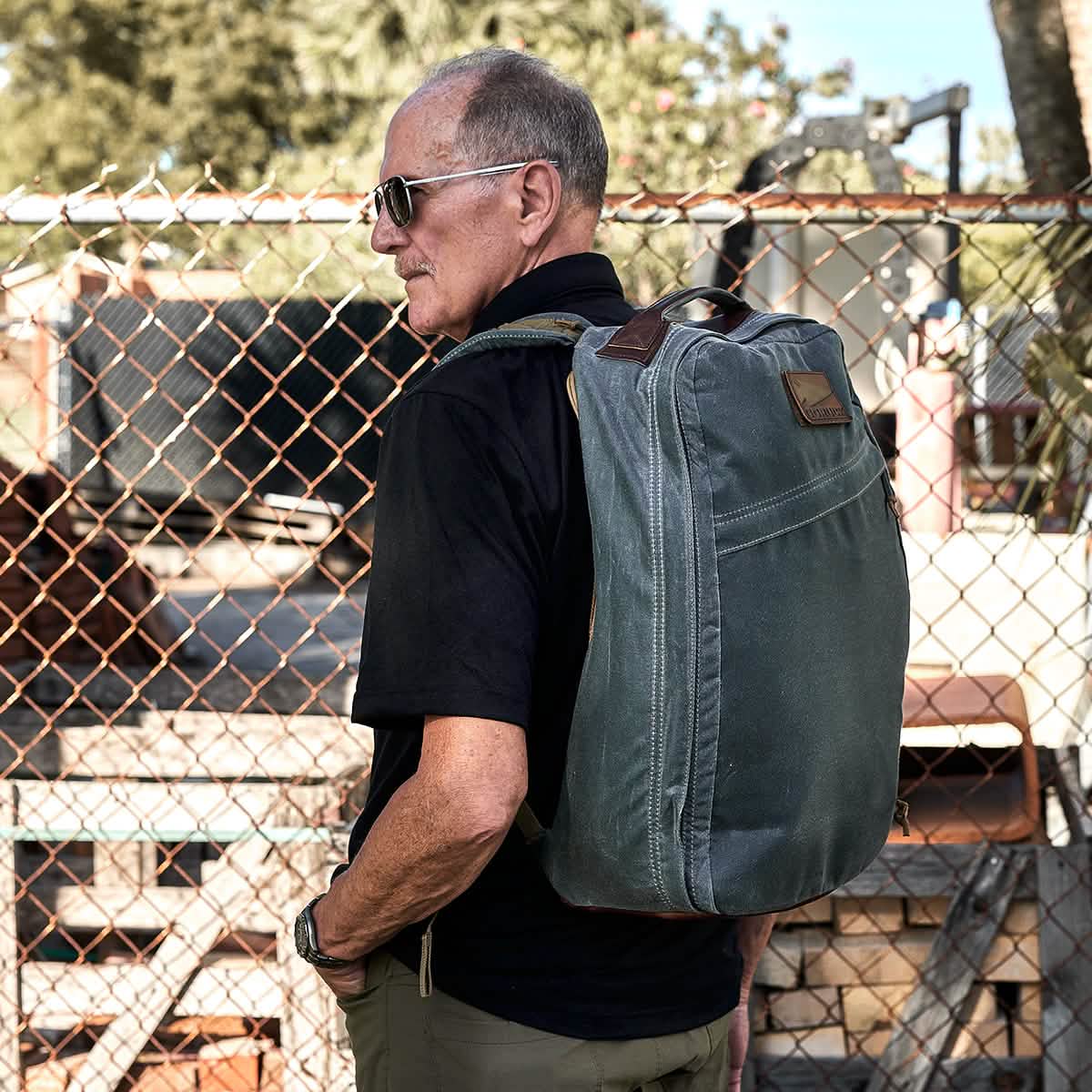 A man wearing sunglasses and a black shirt carries a large green GR1 USA - Heritage rucksack by GORUCK. He is standing in front of a chain-link fence, with industrial equipment and wooden pallets in the background.