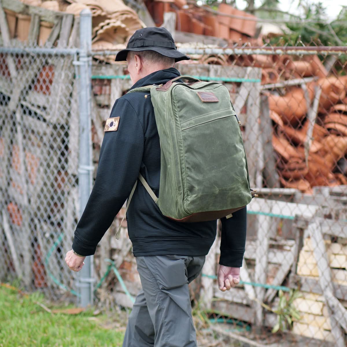 A person wearing a black hat and jacket walks by a fence carrying a large green GR1 USA - Heritage rucksack from GORUCK, crafted from waxed duck canvas. Various materials and terracotta pots are stacked behind the fence.