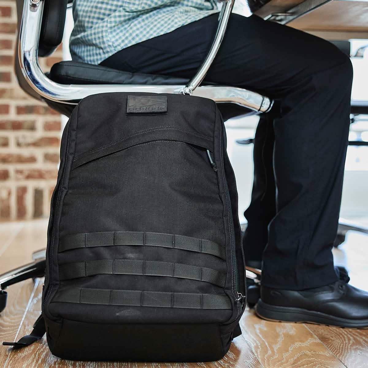 Black GORUCK tactical backpack on wooden floor beside person seated in office chair, brick wall background