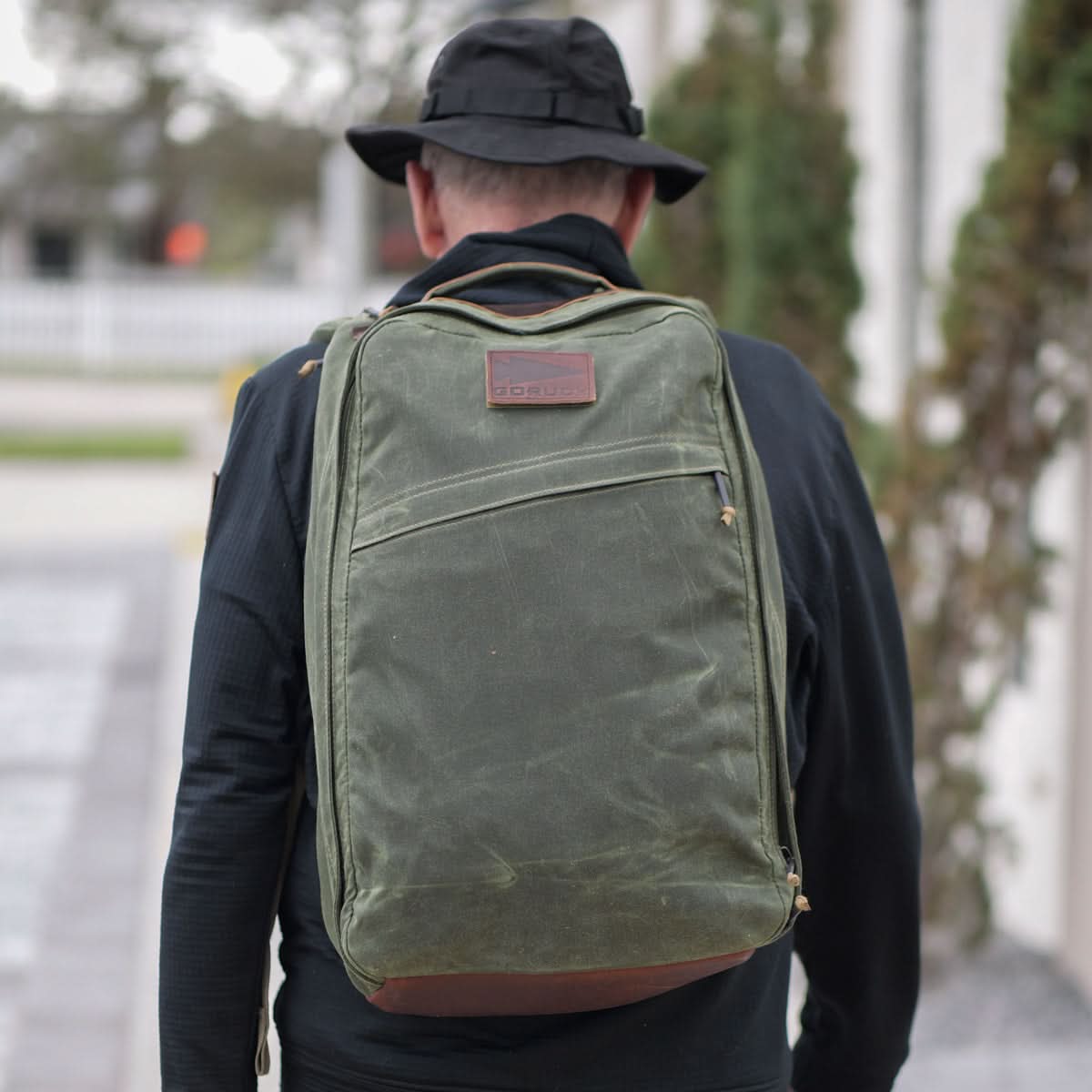 A person wearing a black hat and jacket walks outdoors, carrying a large green GR1 USA - Heritage backpack by GORUCK. The background features a blurred outdoor setting with trees and a building, enhancing the adventurous feel of their journey.