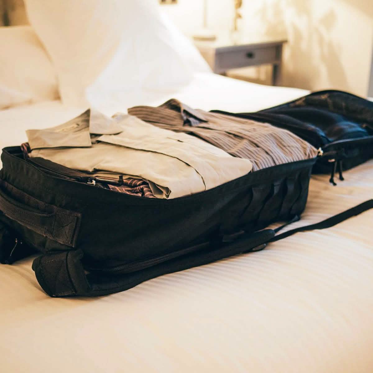 A GR2 travel bag from GORUCK, in sleek black, sits open on a bed, expertly packed with folded clothes. The room is softly lit, and the bed is adorned with white linens. In the background, there is a side table featuring a small lamp.