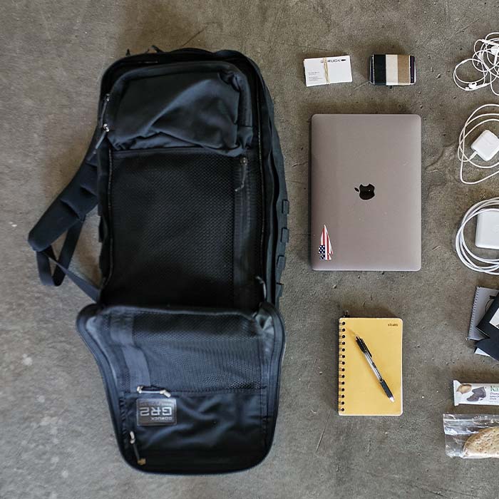 Open black backpack next to a laptop, notebook, pen, chargers, cords, and small organized items on the floor.