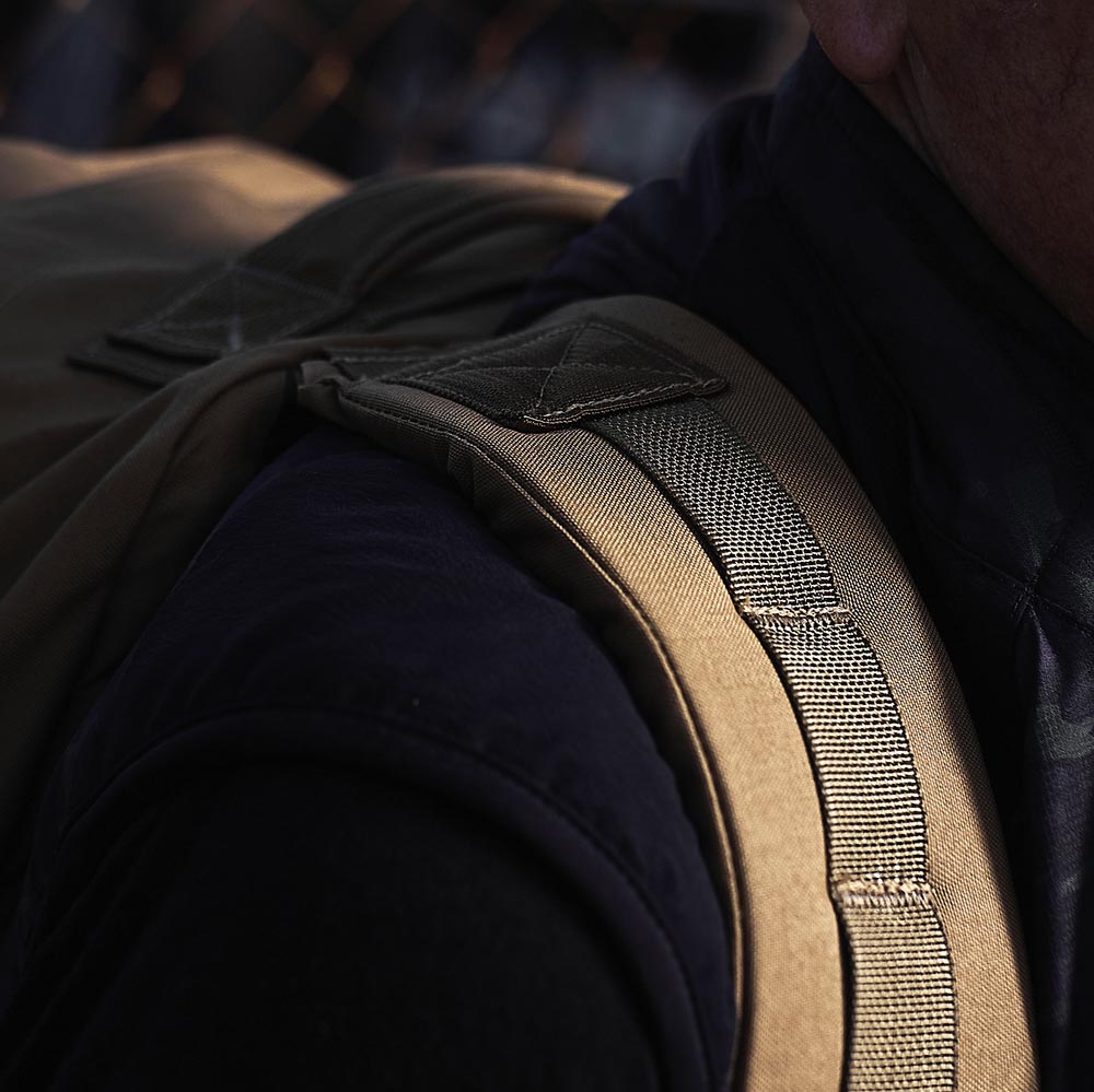 Close-up of a tan backpack strap on a person's shoulder, with part of their dark shirt and vest visible.