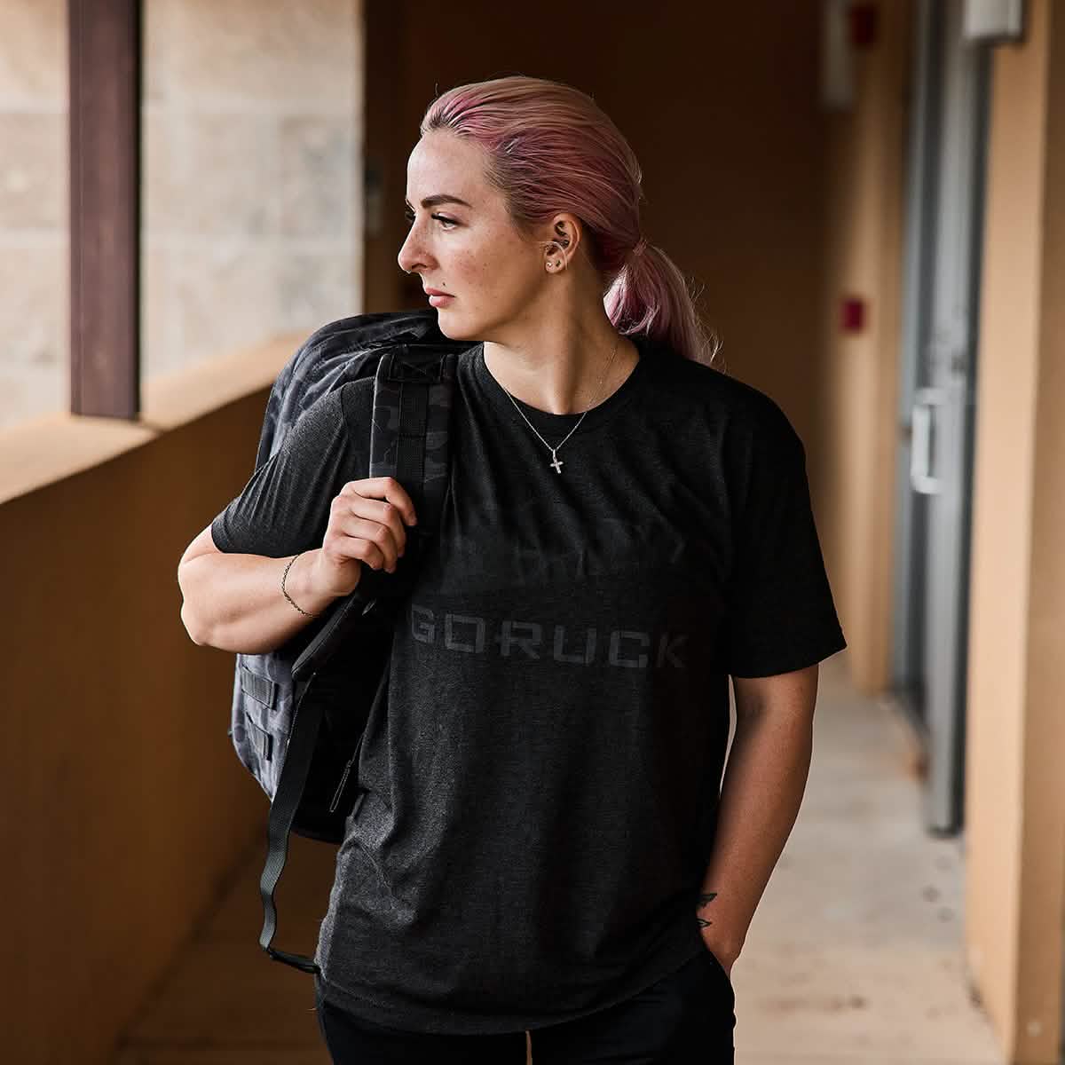 A person with pink hair stands in a hallway, glancing to the side. They wear a dark t-shirt and effortlessly carry a GORUCK GR2 ruck over one shoulder.