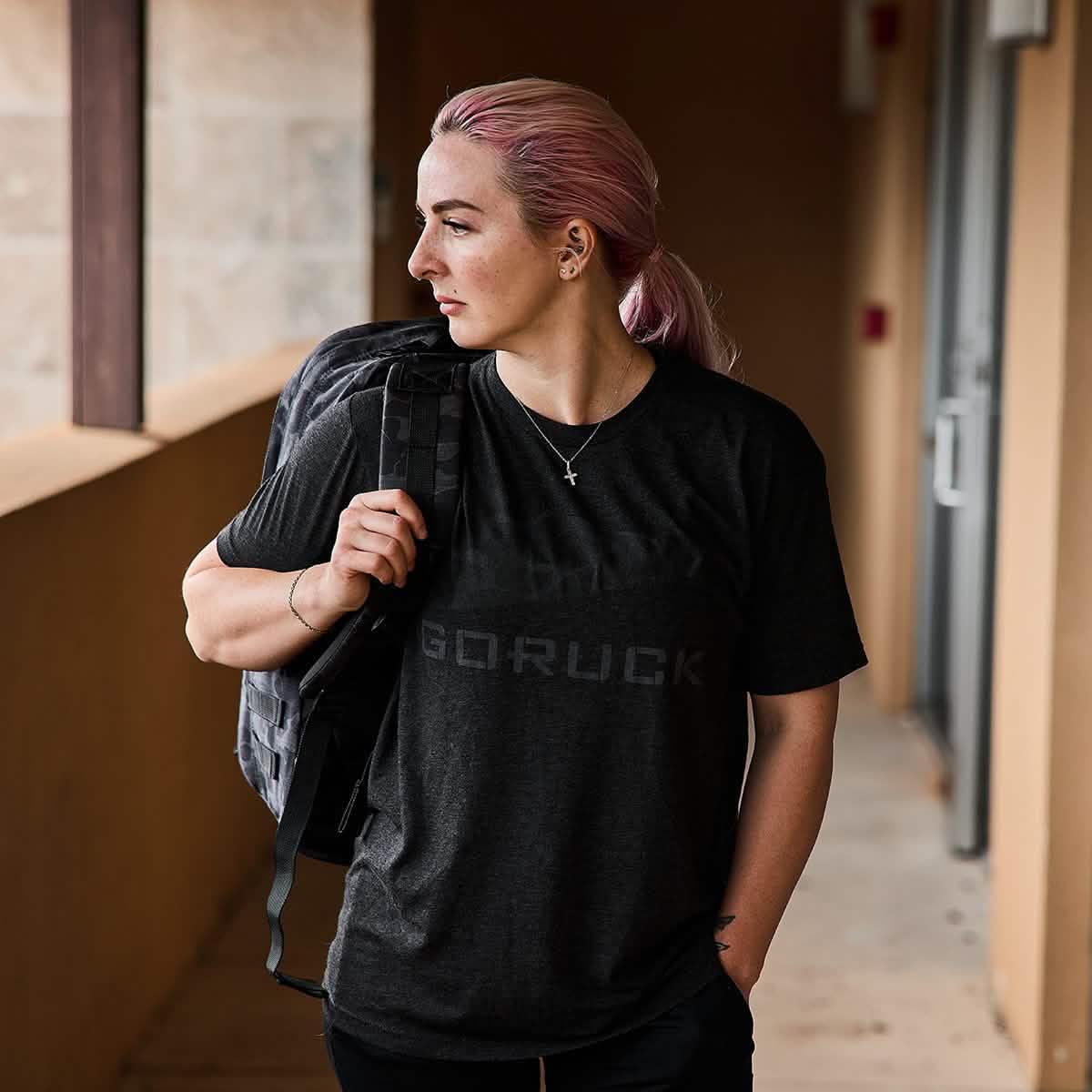 Woman with pink hair wearing black GORUCK t-shirt and carrying a black rucksack on shoulder in corridor