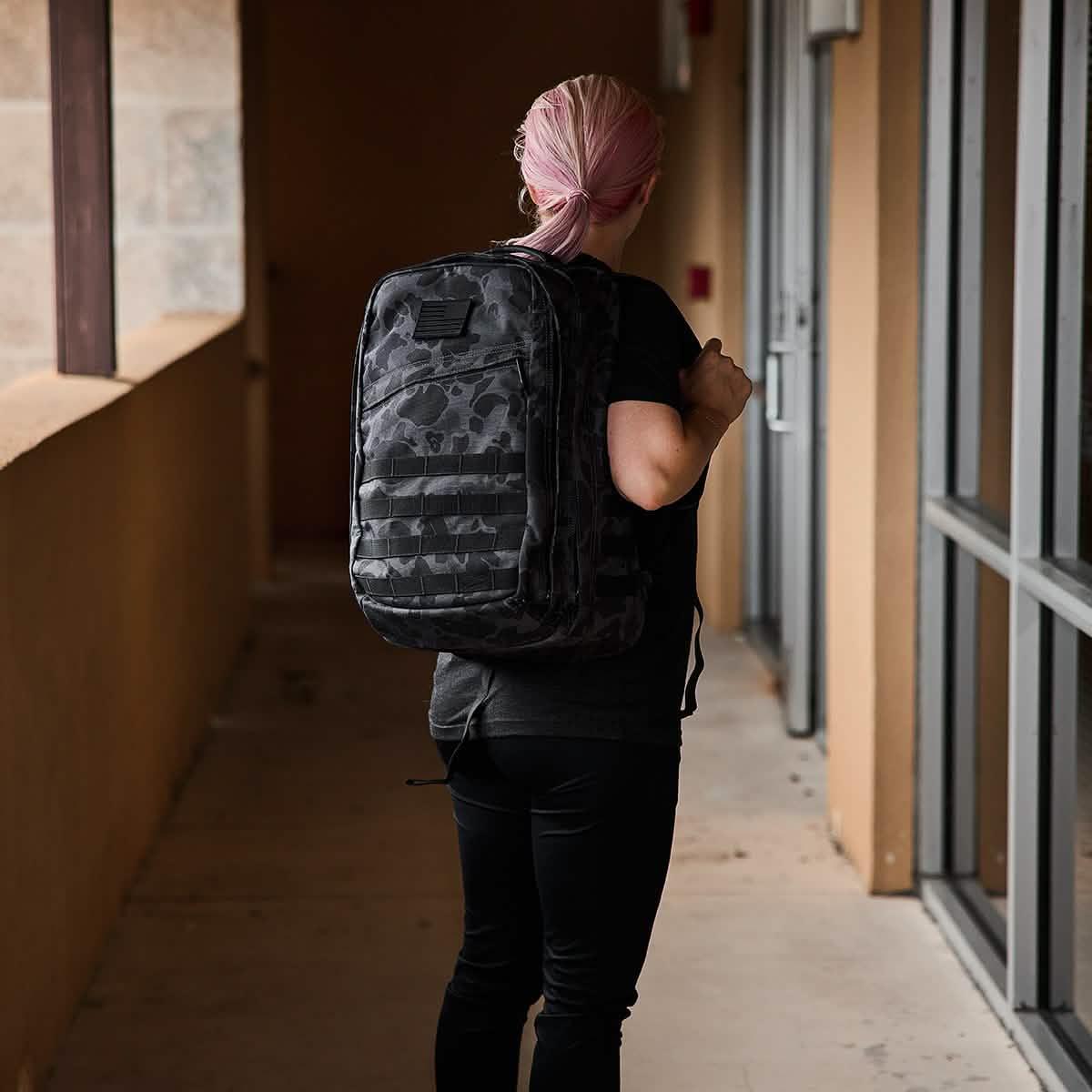 Person with pink hair wearing black clothes carrying a GORUCK black Frogskin tactical backpack in a corridor