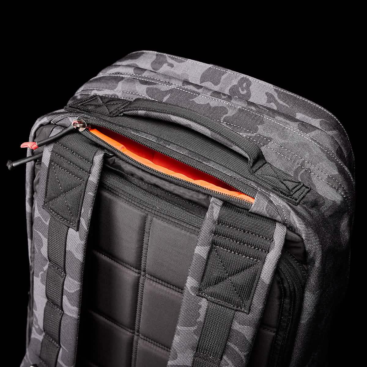 A close-up image of the GR2 backpack by GORUCK, showcasing its gray and black camouflage pattern reminiscent of those employed by Special Forces, set against a black background. The backpack features padded black straps and a partially open orange zipper revealing the bright orange interior lining.
