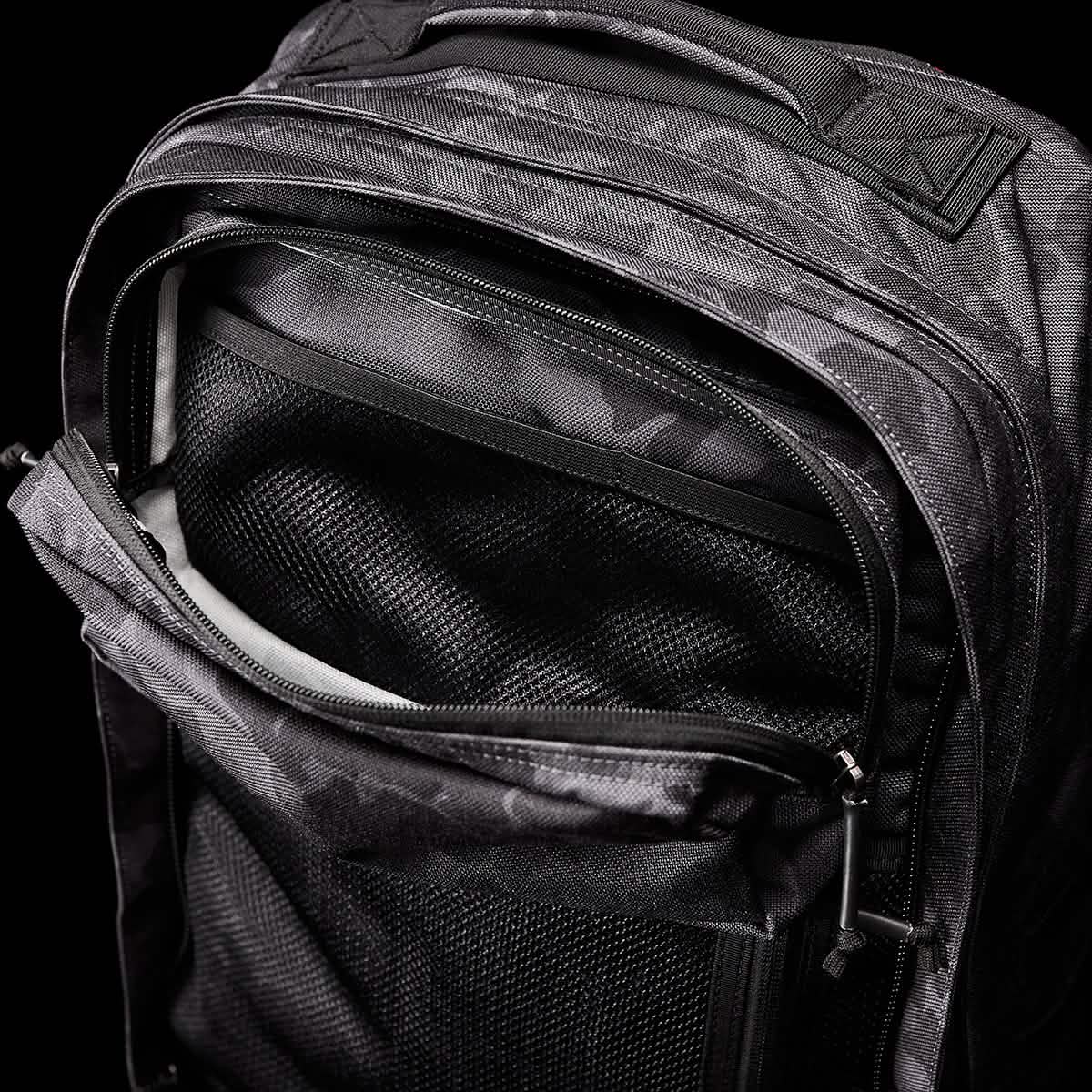 A close-up of a black GORUCK GR2 backpack reveals an open front pocket with visible mesh lining, similar in style to a Special Forces ruck. The zippers and fabric texture stand out prominently against the black background.
