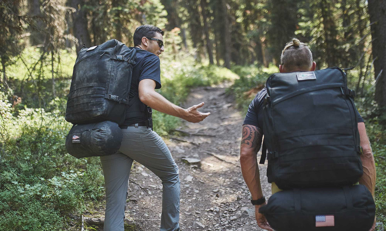 Two people with large backpacks hiking up a dirt trail through a forested area, talking to each other.