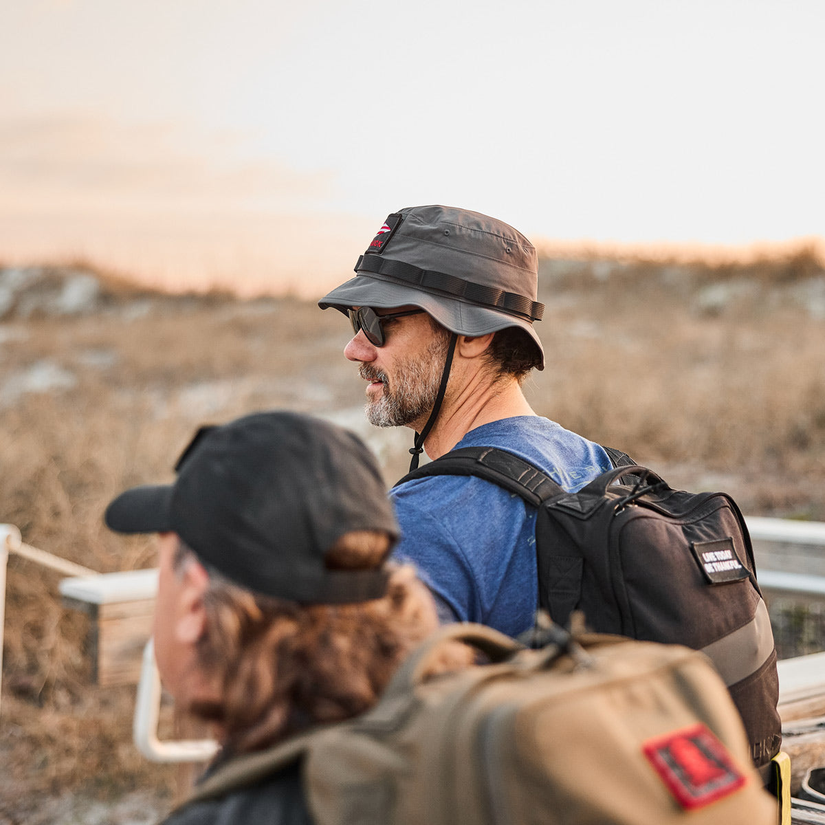 Two men wearing hats and backpacks walk outdoors near dry grass under a bright sky.