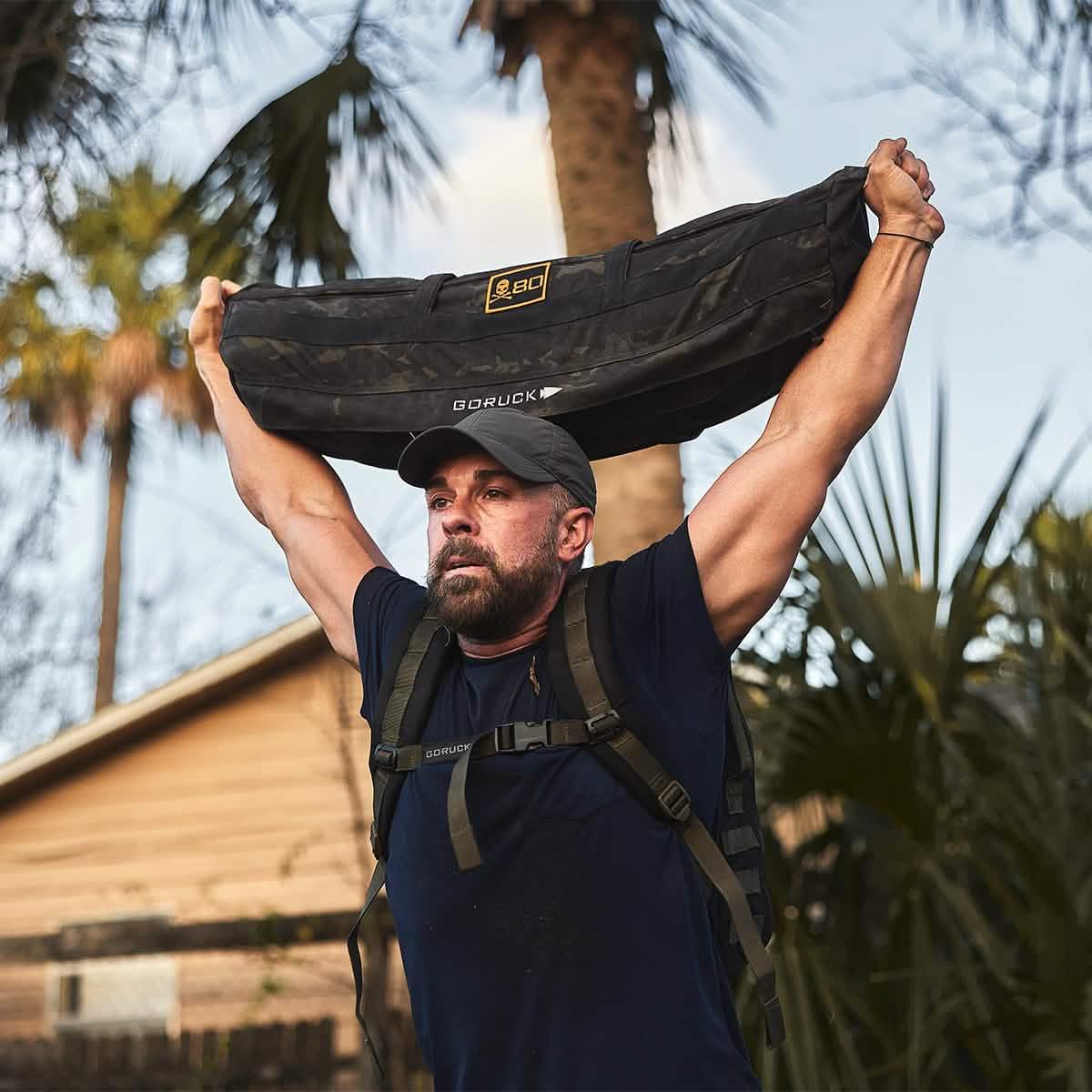 Man outdoors lifting GORUCK sandbag overhead, wearing rucking gear and GORUCK cap.