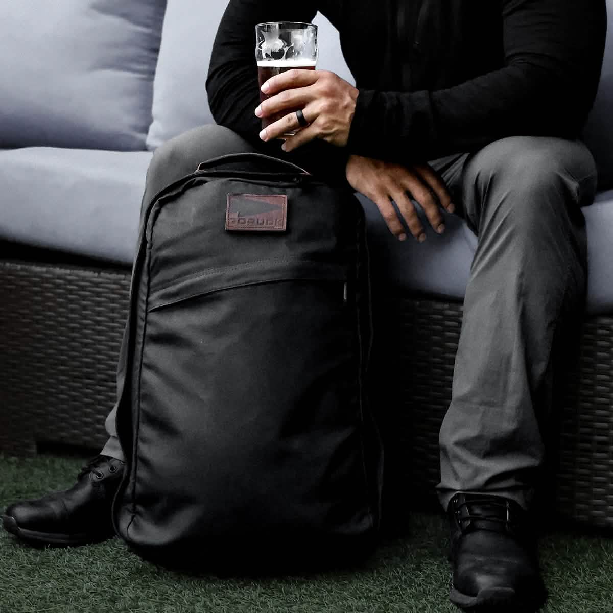 A person sitting on a couch outdoors, holding a glass of beer in one hand and resting the other on a GR1 USA - Heritage rucksack by GORUCK placed on the ground. The person is wearing dark clothing, creating a relaxed atmosphere.