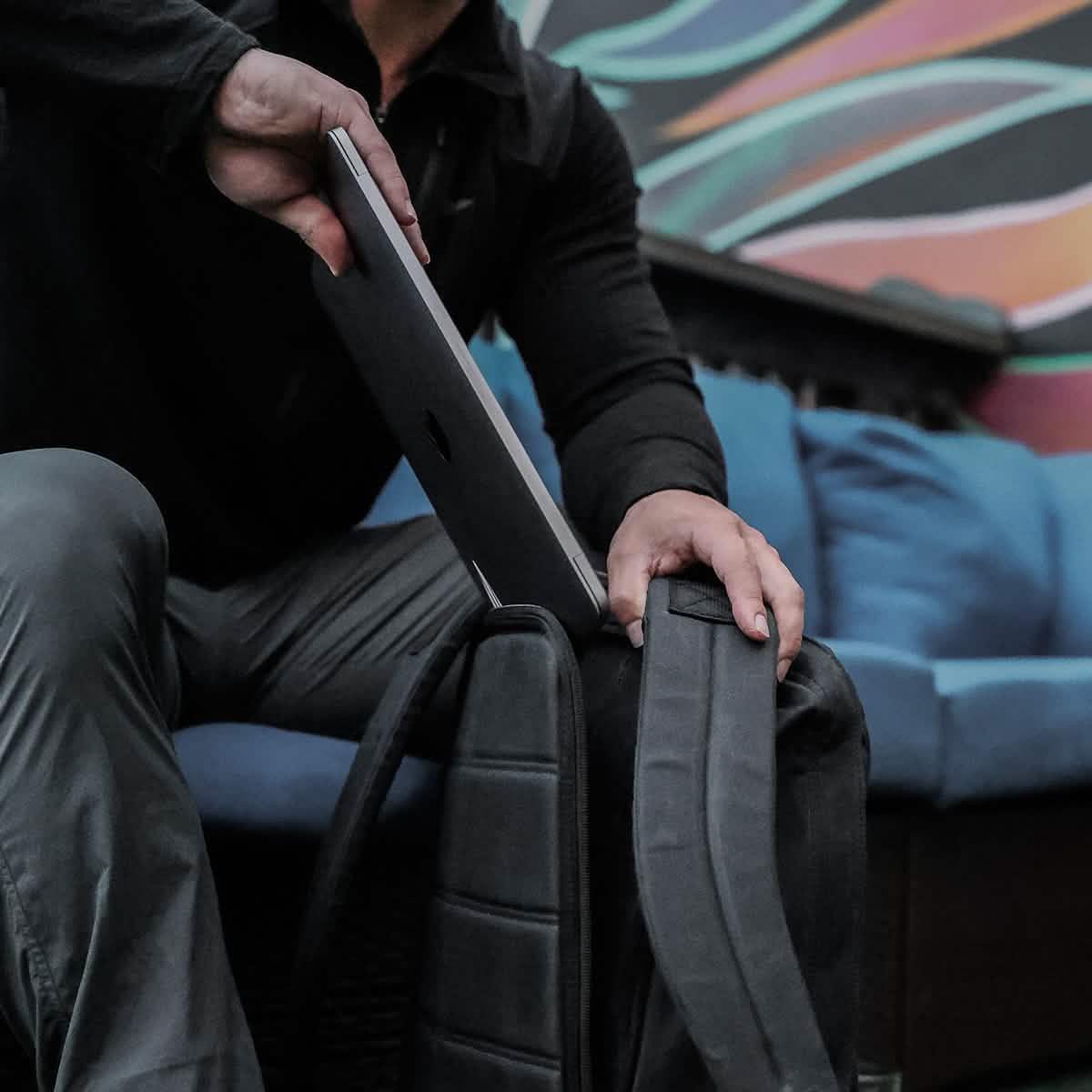 Person placing laptop into black GORUCK backpack with blue couch and colorful wall art in background