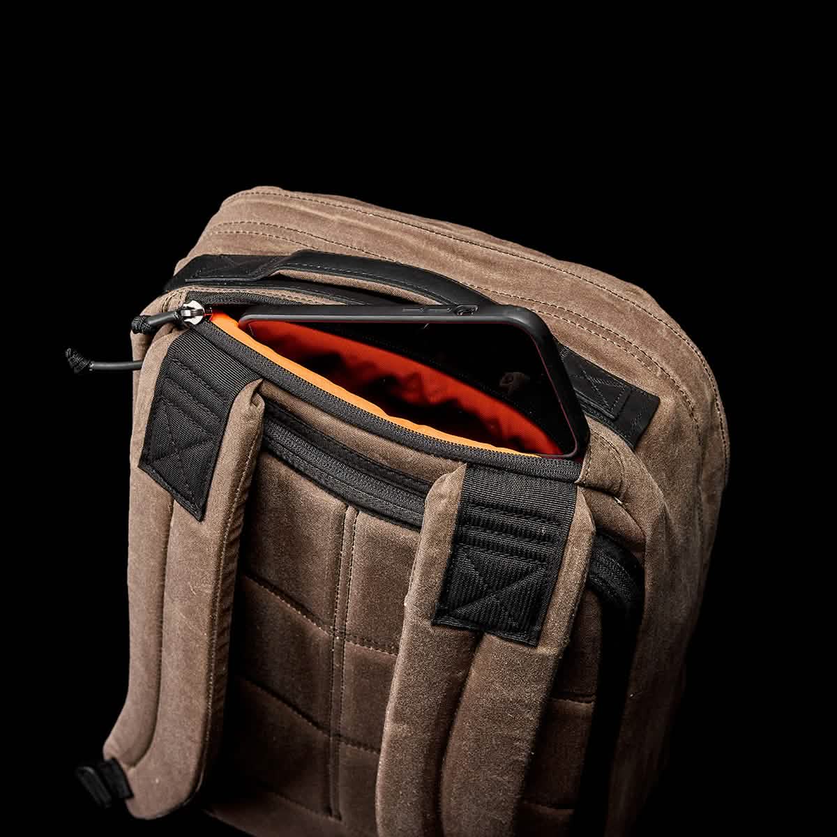The GR1 USA - Heritage from GORUCK is showcased against a black background, with its top zipper open to display an orange interior and a smartphone peeking out. Made from waxed duck canvas, this rucksack features black straps and stitching.
