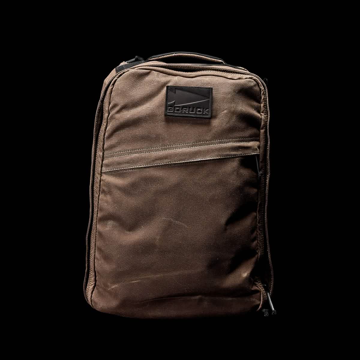 A brown GR1 USA - Heritage backpack from GORUCK is centered against a black background. It features a sturdy, rectangular design with a visible front zipper pocket and a GORUCK logo patch at the top.