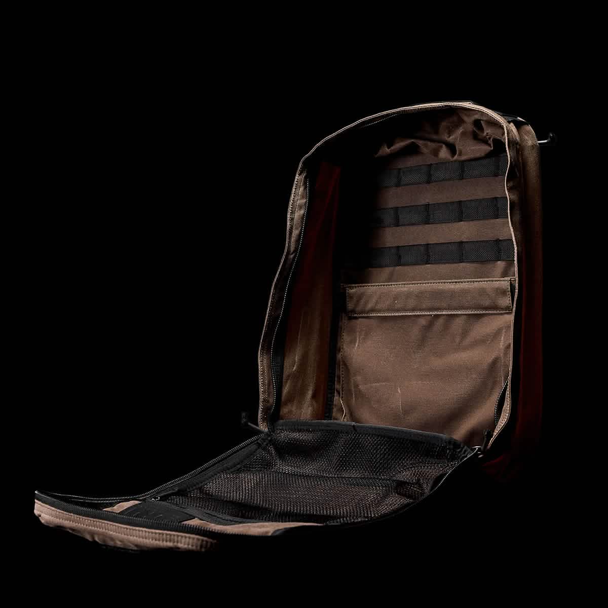 The GR1 USA - Heritage by GORUCK is pictured open against a black background, revealing its tan interior with organized pockets and compartments. Made from waxed duck canvas, it boasts a utilitarian and functional design with mesh sections and horizontal loops for attachments.