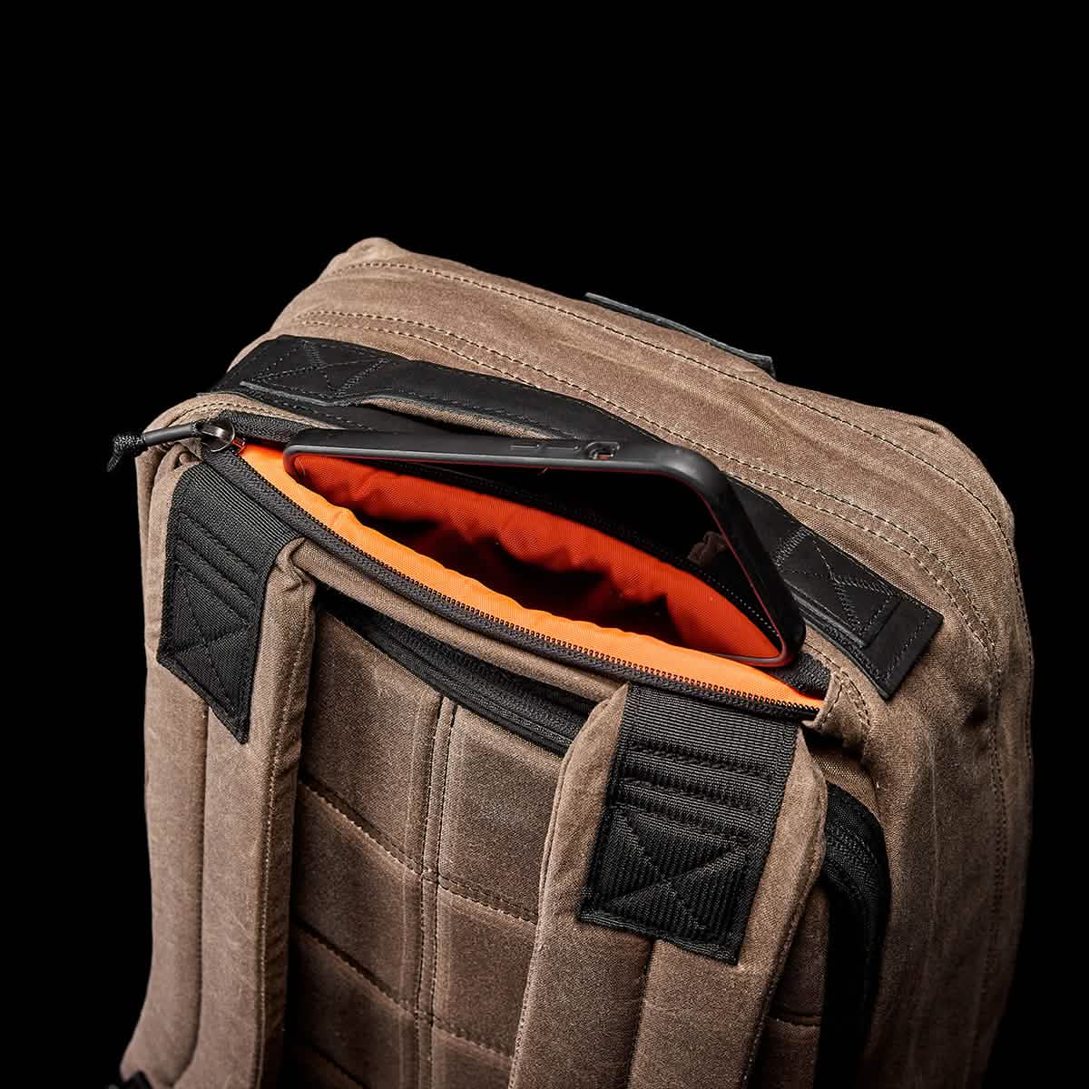 A close-up shot features a brown GR1 USA - Heritage by GORUCK, showcasing its black straps and partially open design that reveals a tablet inside. The interior lining is vibrant orange, providing contrast to the rugged exterior made from waxed duck canvas. The background is solid black.