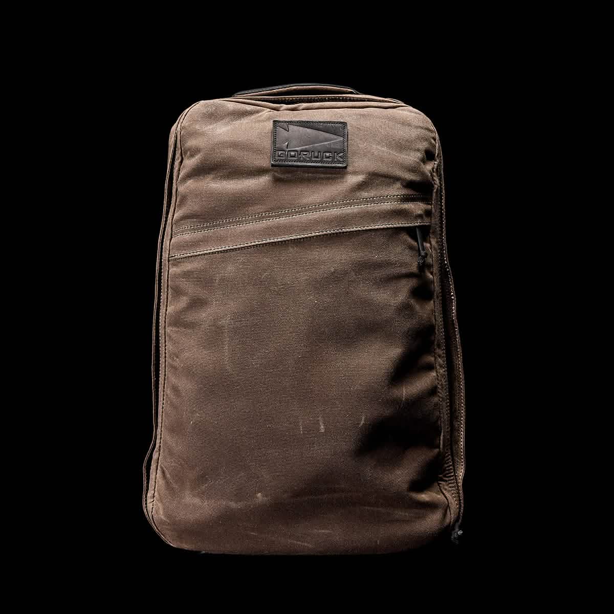 The GR1 USA - Heritage by GORUCK is a brown retro canvas rucksack showcasing a rectangular patch with an embossed logo, set against a black background. It features a front zipper pocket and seems to be crafted from durable fabric.