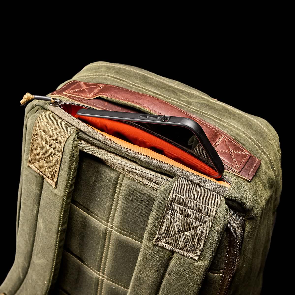 A GORUCK GR1 USA - Heritage rucksack with quilted stitching is unzipped, revealing an orange interior and a black smartphone partially visible inside the pocket against a black background.