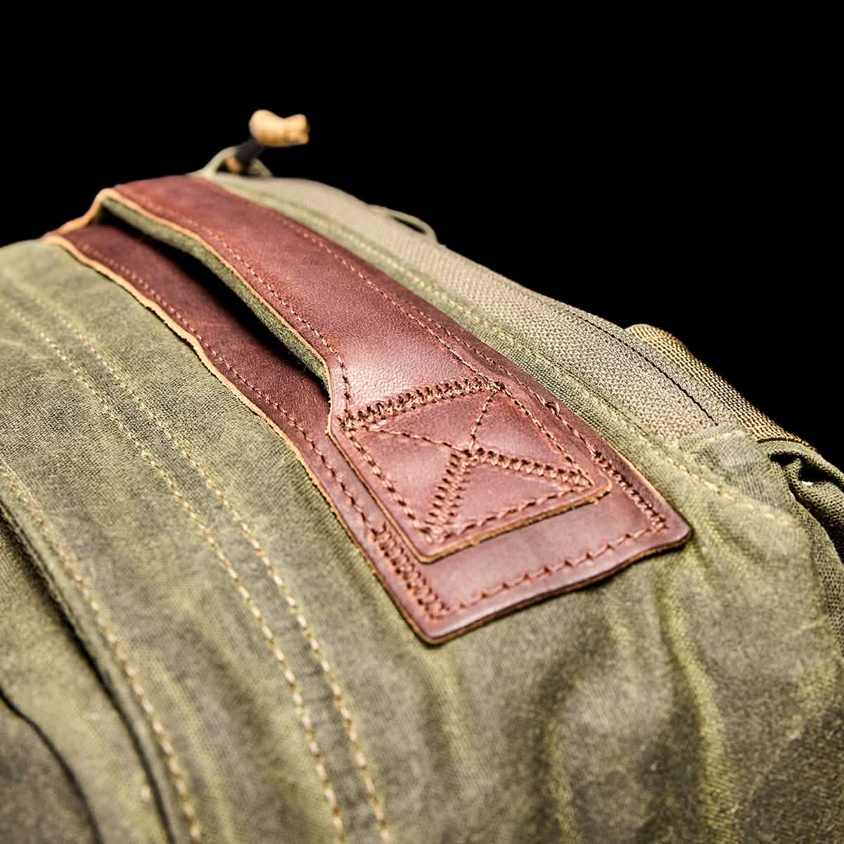 Close-up of the GORUCK GR1 USA - Heritage rucksack showcasing its retro design with a brown leather handle and intricate stitching details. The focus is on the waxed duck canvas texture and craftsmanship, set against a black background.