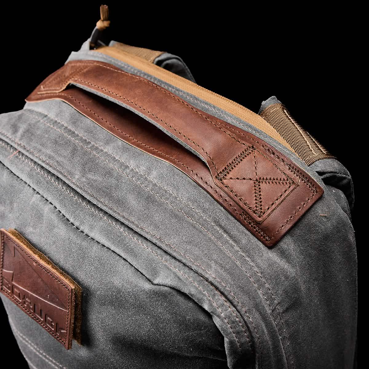 Close-up of a rugged gray GR1 USA - Heritage rucksack from GORUCK featuring a brown leather handle and tan accents. The waxed canvas texture and precise stitching enhance its durability. The black background emphasizes the backpack’s detailed design.