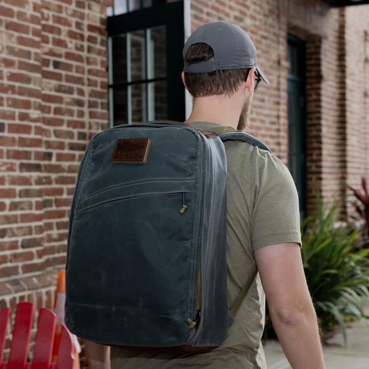 Man wearing a gray GORUCK backpack with a leather patch walking in front of a brick building